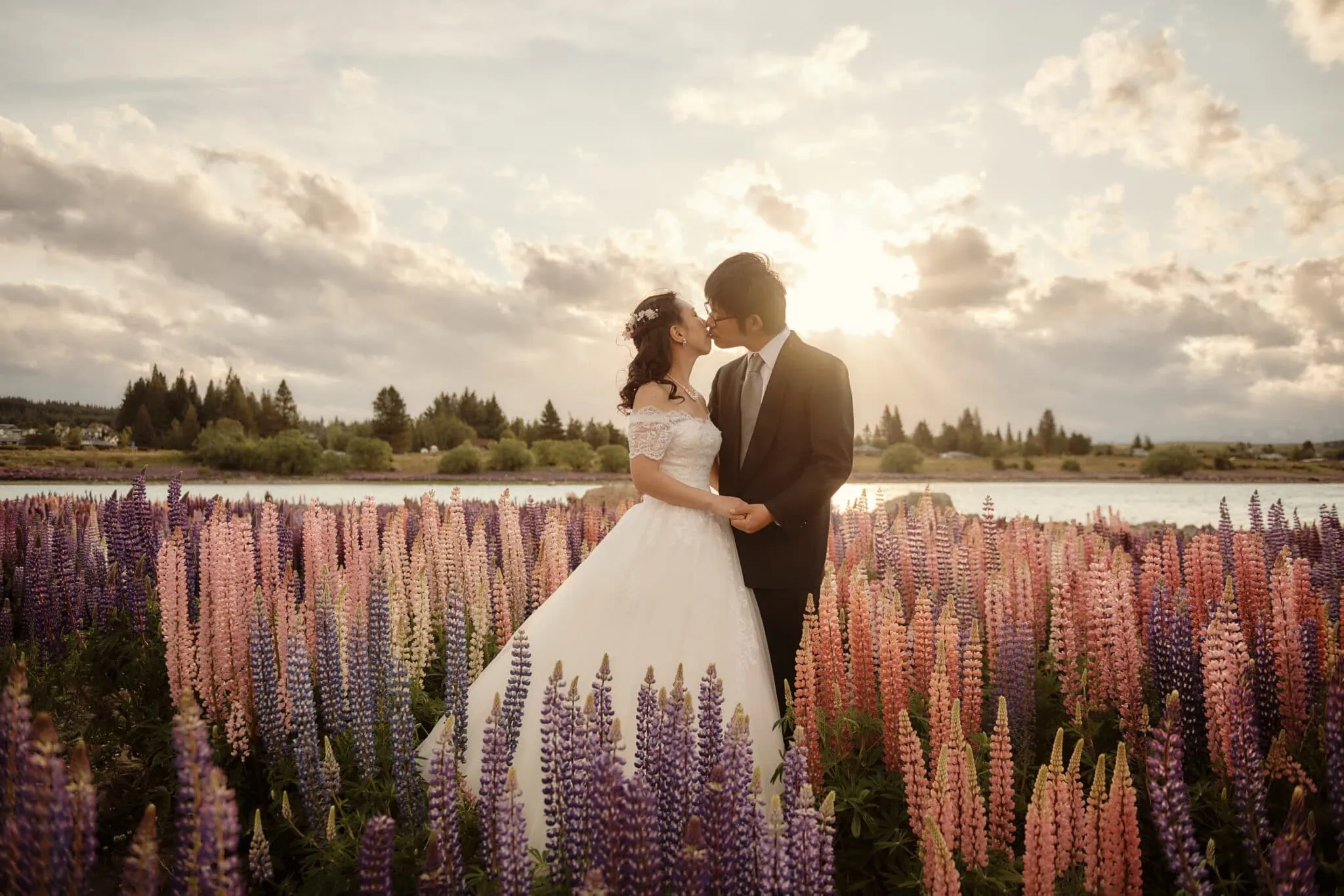 queenstown new zealand heli wedding elopement photographer videographer | A couple, Eri & Youhei, dressed in wedding attire stands in a field of colorful flowers, kissing at sunset with a lake and trees in the background—a beautiful moment captured by Queenstown Pre-wedding Photography.