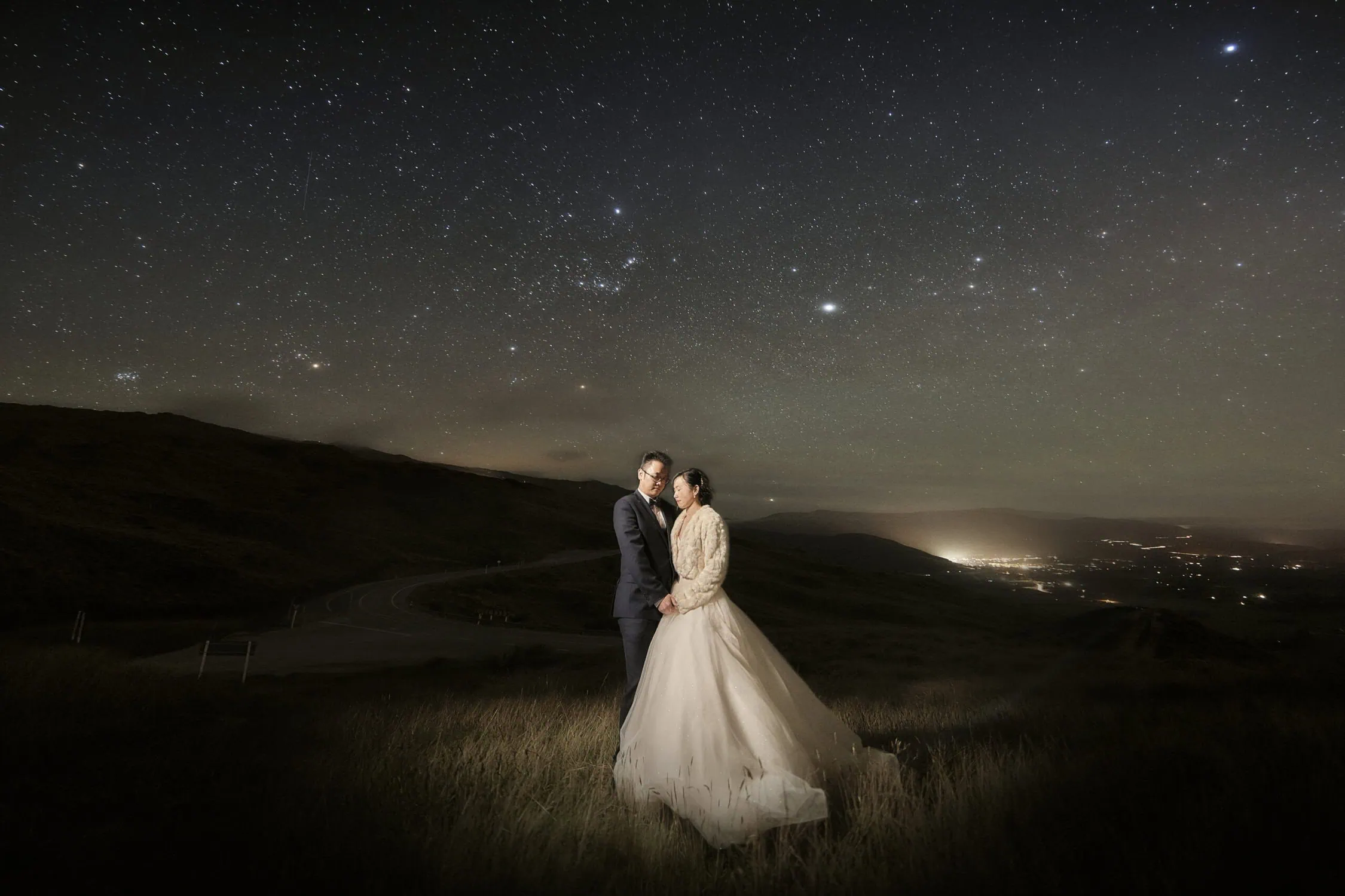 queenstown new zealand heli wedding elopement photographer videographer | A couple in wedding attire stands under a starry night sky in a grassy landscape with hills and city lights in the distance, capturing the essence of Queenstown Pre-wedding Photography.