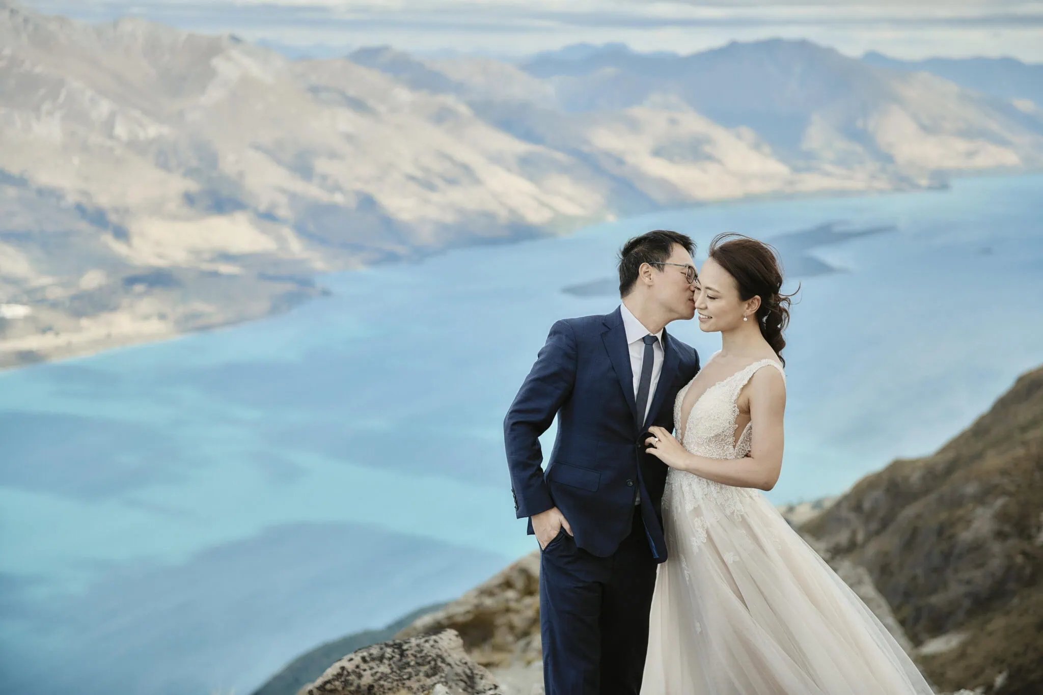 queenstown new zealand heli wedding elopement photographer videographer | A couple in formal attire stands close together on a mountainside overlooking a large lake and distant mountains. During their Queenstown Pre-Wedding shoot, the man in a suit kisses Mindy’s forehead as they share an intimate moment.