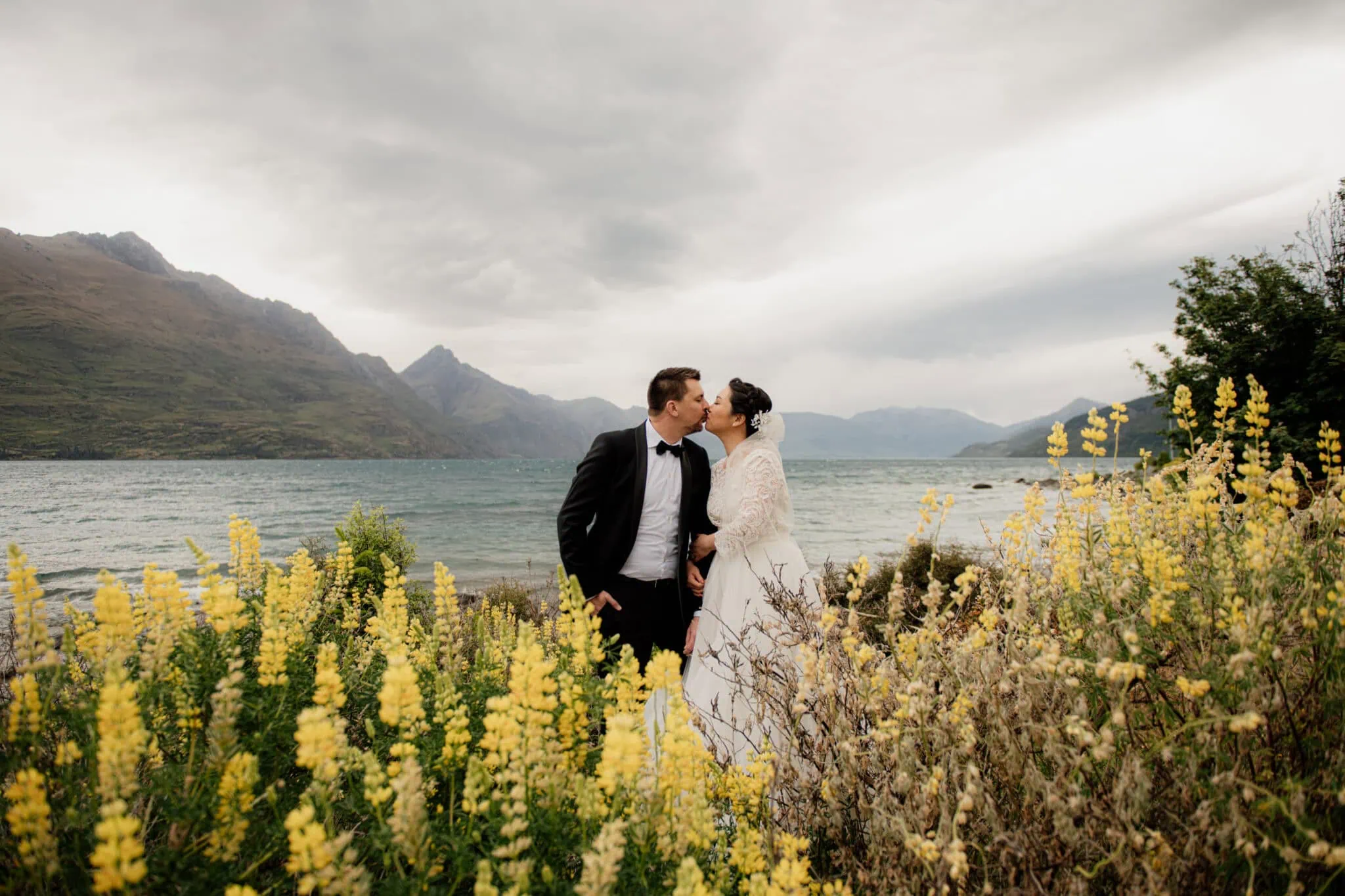 queenstown new zealand heli wedding elopement photographer videographer | A couple in formal attire kisses by a lake surrounded by yellow flowers, with mountains in the background under a cloudy sky, capturing the essence of Queenstown pre-wedding photography.