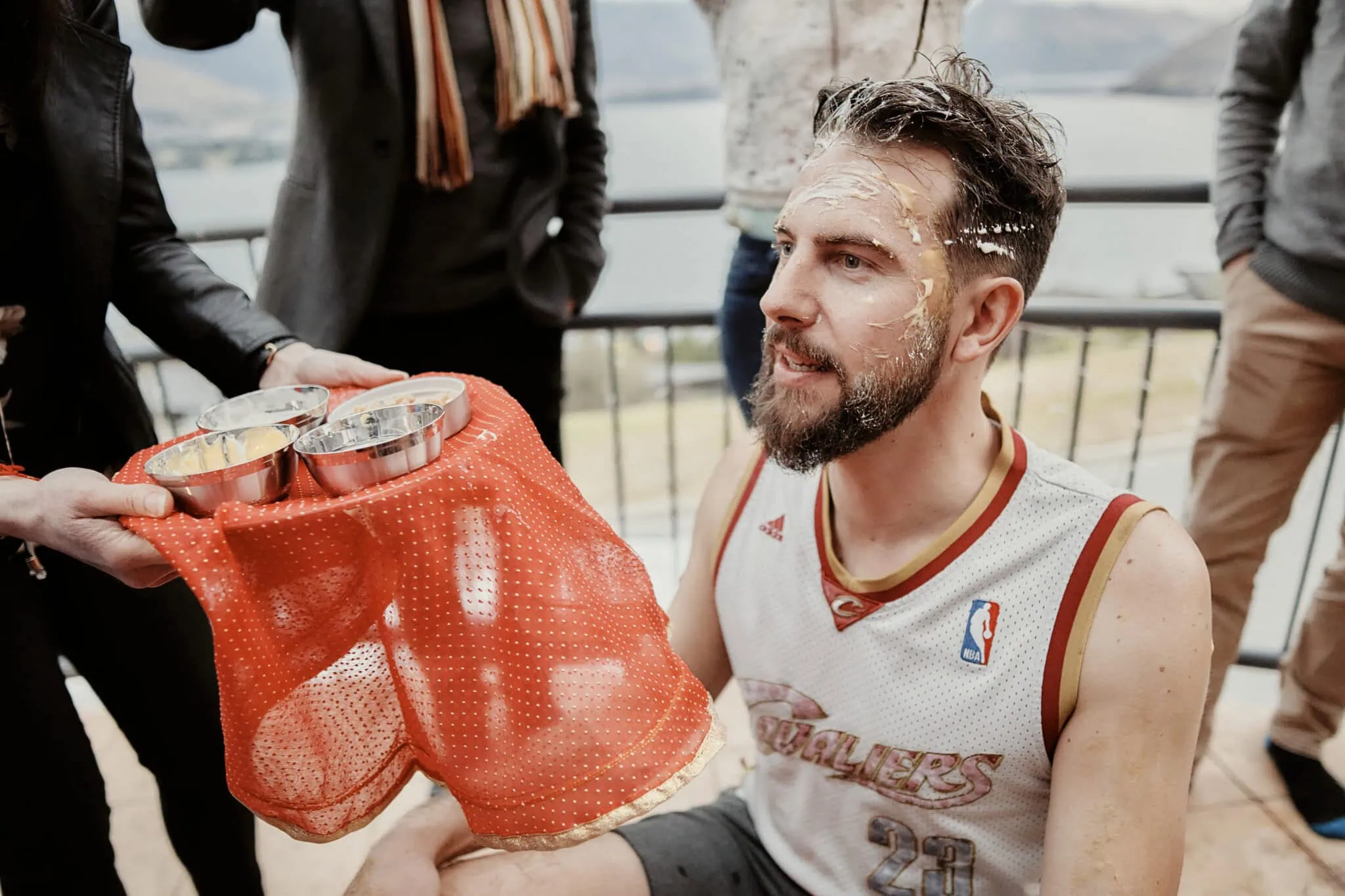 queenstown new zealand heli wedding elopement photographer videographer | In a humorous scene from Jasveen & Ben's Queenstown elopement, a man in a basketball jersey sits with food on his face, while another person holds a tray of food-covered metal cups partially obscured by mesh fabric. Others stand around in the background, capturing the candid moment.