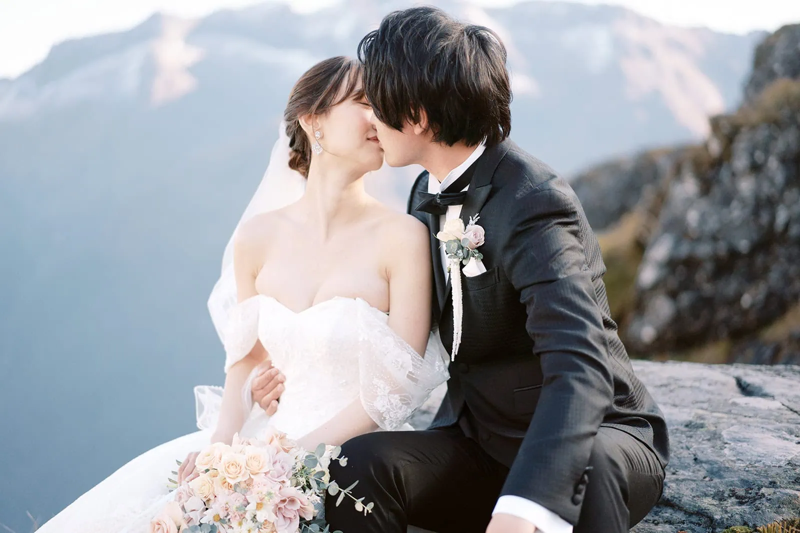 queenstown new zealand heli wedding elopement photographer videographer | A bride and groom in wedding attire share a kiss while seated on a rock, with a mountainous landscape in the background. The bride holds a bouquet of flowers, capturing the essence of Queenstown elopement in this stunning piece of wedding photography.
