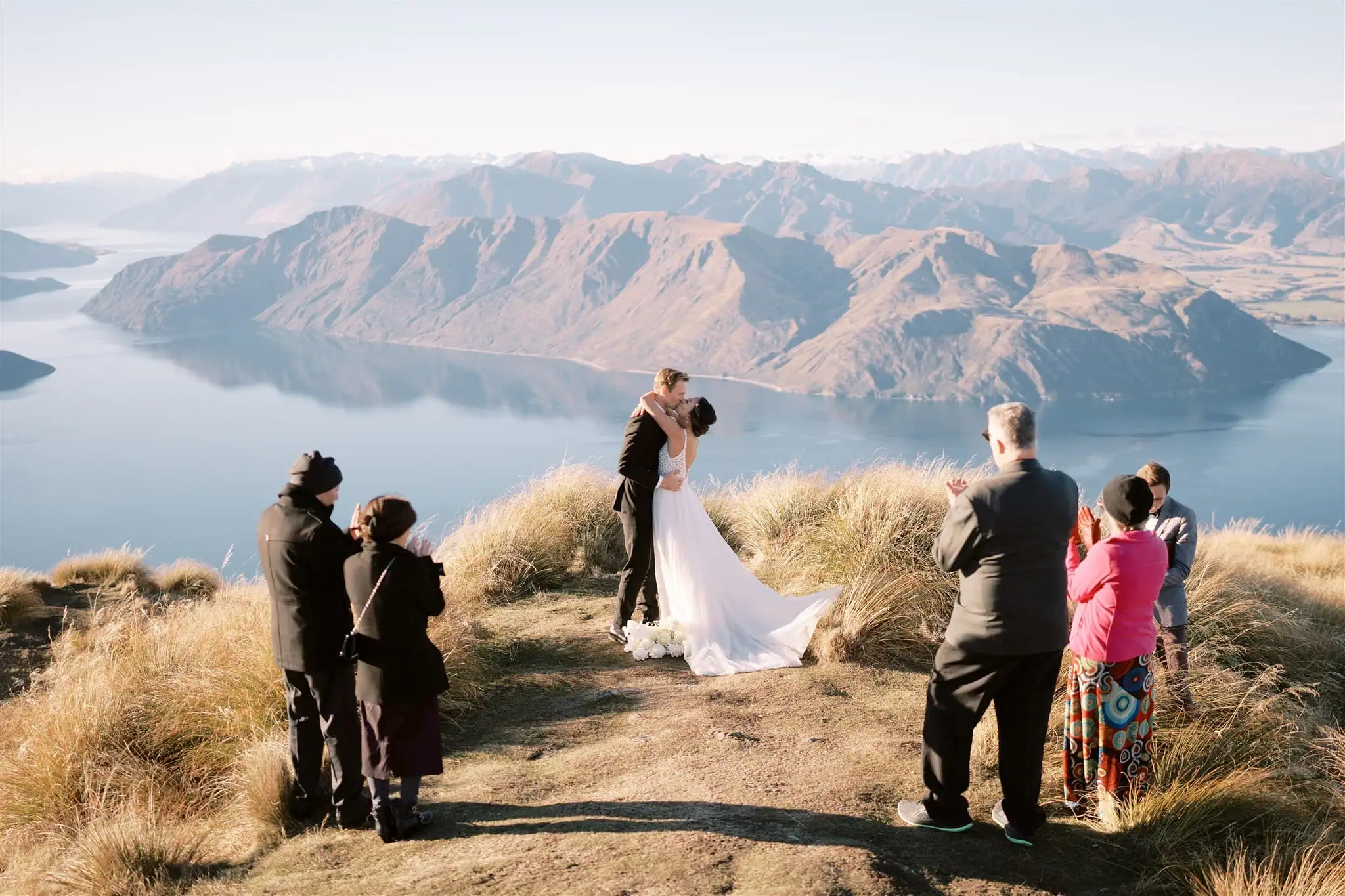 queenstown new zealand heli wedding elopement photographer videographer | A couple kisses at their elopement wedding on a mountaintop in Queenstown, with a scenic view of the lake and mountains, surrounded by six guests. The photography captures their intimate outdoor ceremony perfectly.