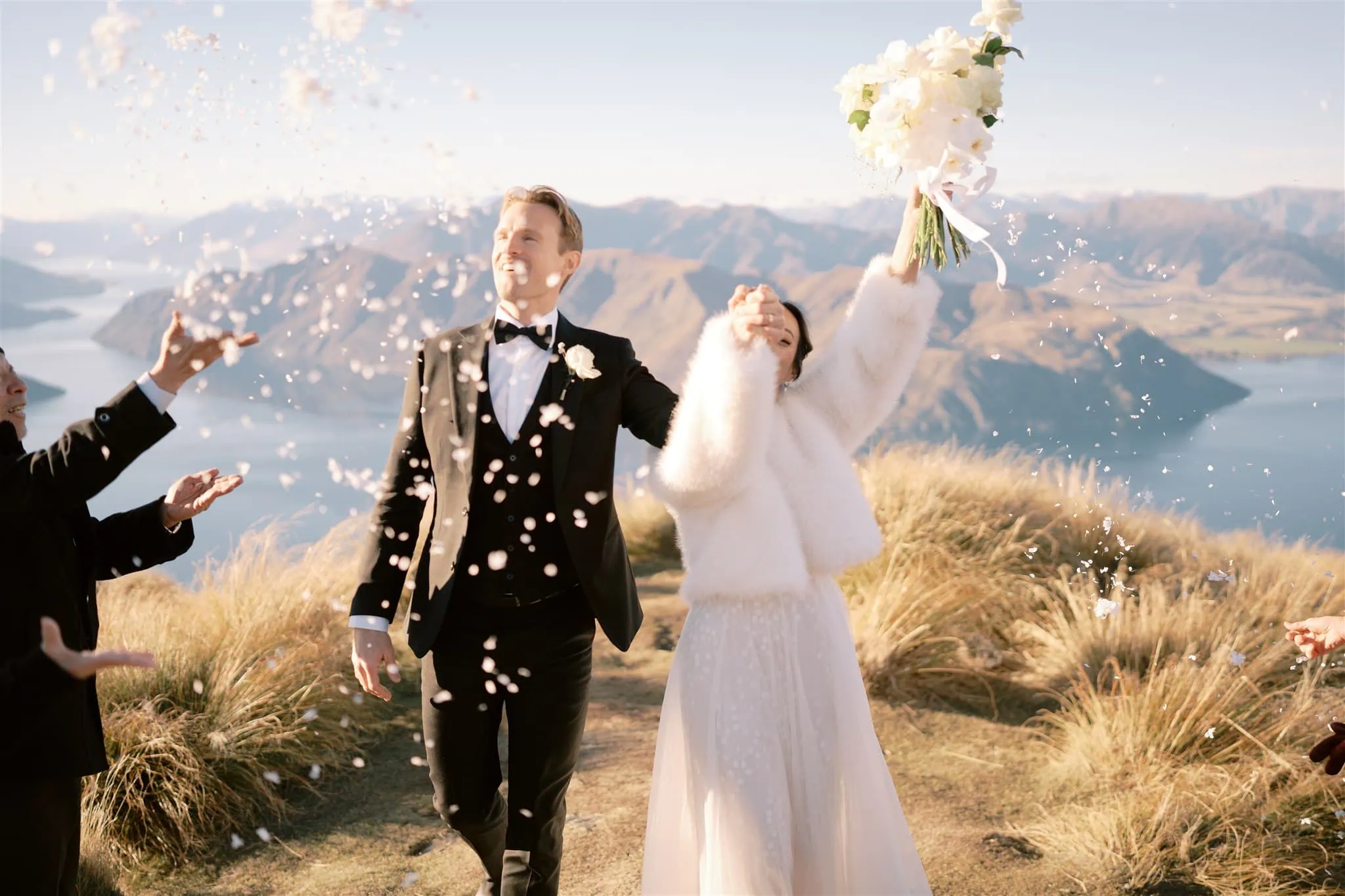 queenstown new zealand heli wedding elopement photographer videographer | Tracey & Sam's Queenstown elopement captures a dreamy moment as they celebrate outdoors, surrounded by mountains and lakes. Confetti flies as the groom in his black suit and the bride in her white dress and fur jacket, holding a bouquet, embrace their new journey. Perfect for wedding photography enthusiasts.