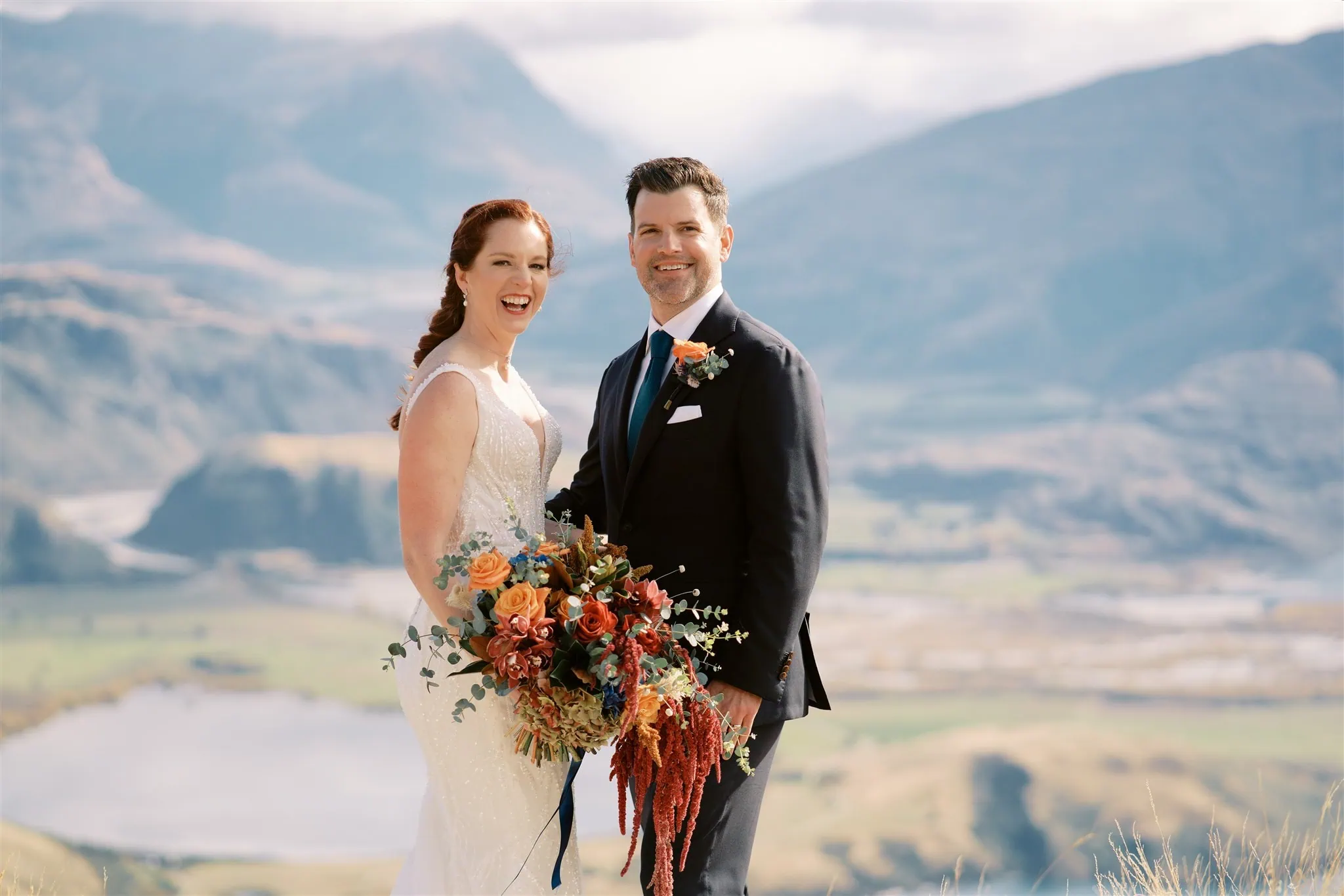 queenstown new zealand heli wedding elopement photographer videographer | A bride and groom stand outdoors, smiling in front of the breathtaking mountainous landscape of Queenstown. The bride holds a bouquet of flowers as they pose in their elegant wedding attire, capturing the perfect moment with exquisite wedding photography from their enchanting elopement.
