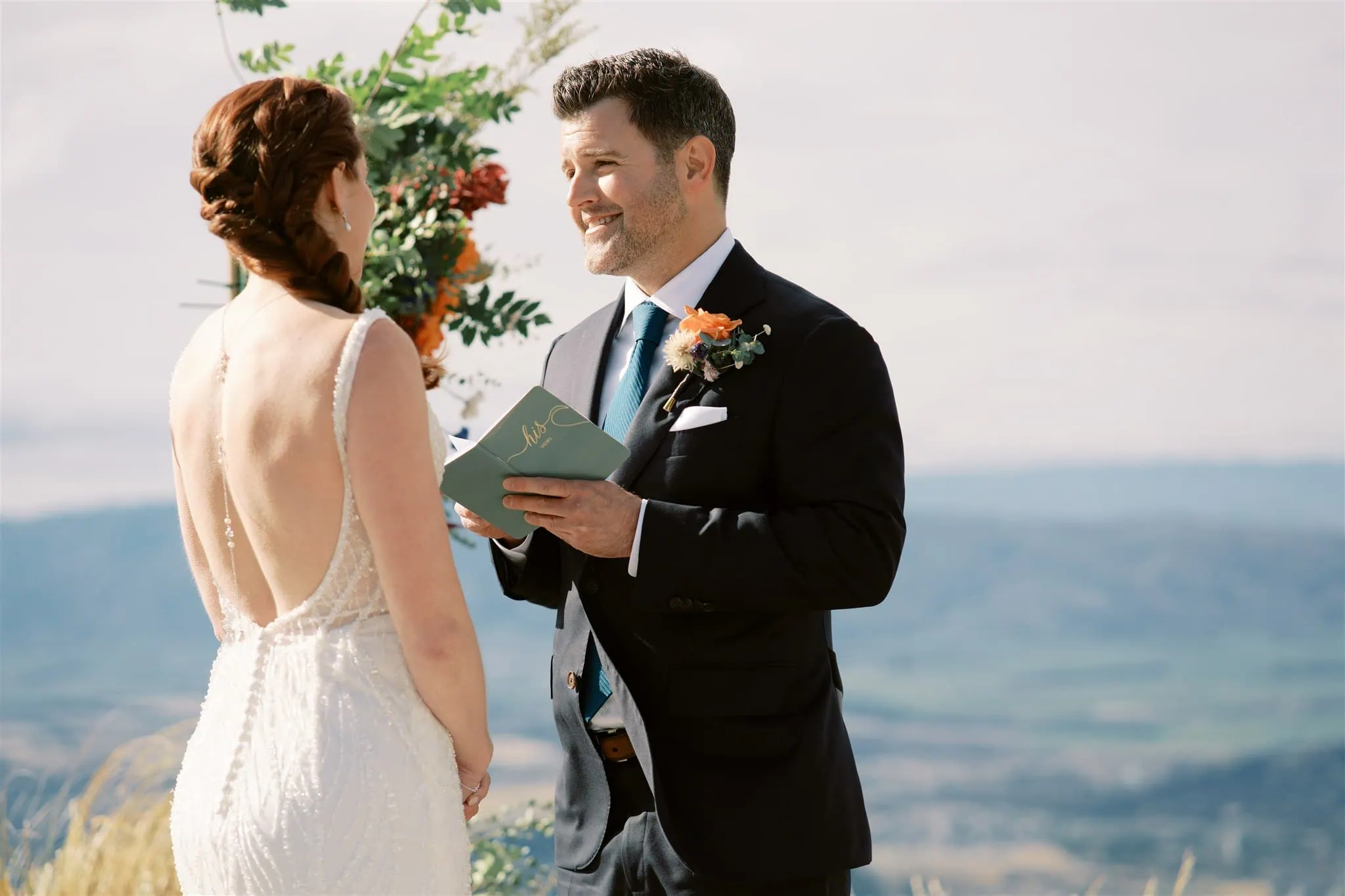 queenstown new zealand heli wedding elopement photographer videographer | A bride and groom, Sarah & John, stand outdoors during their Queenstown elopement. The groom is holding a book and reading, while the bride faces him. In the background, there are mountains and a partly cloudy sky—perfect for wedding photography.