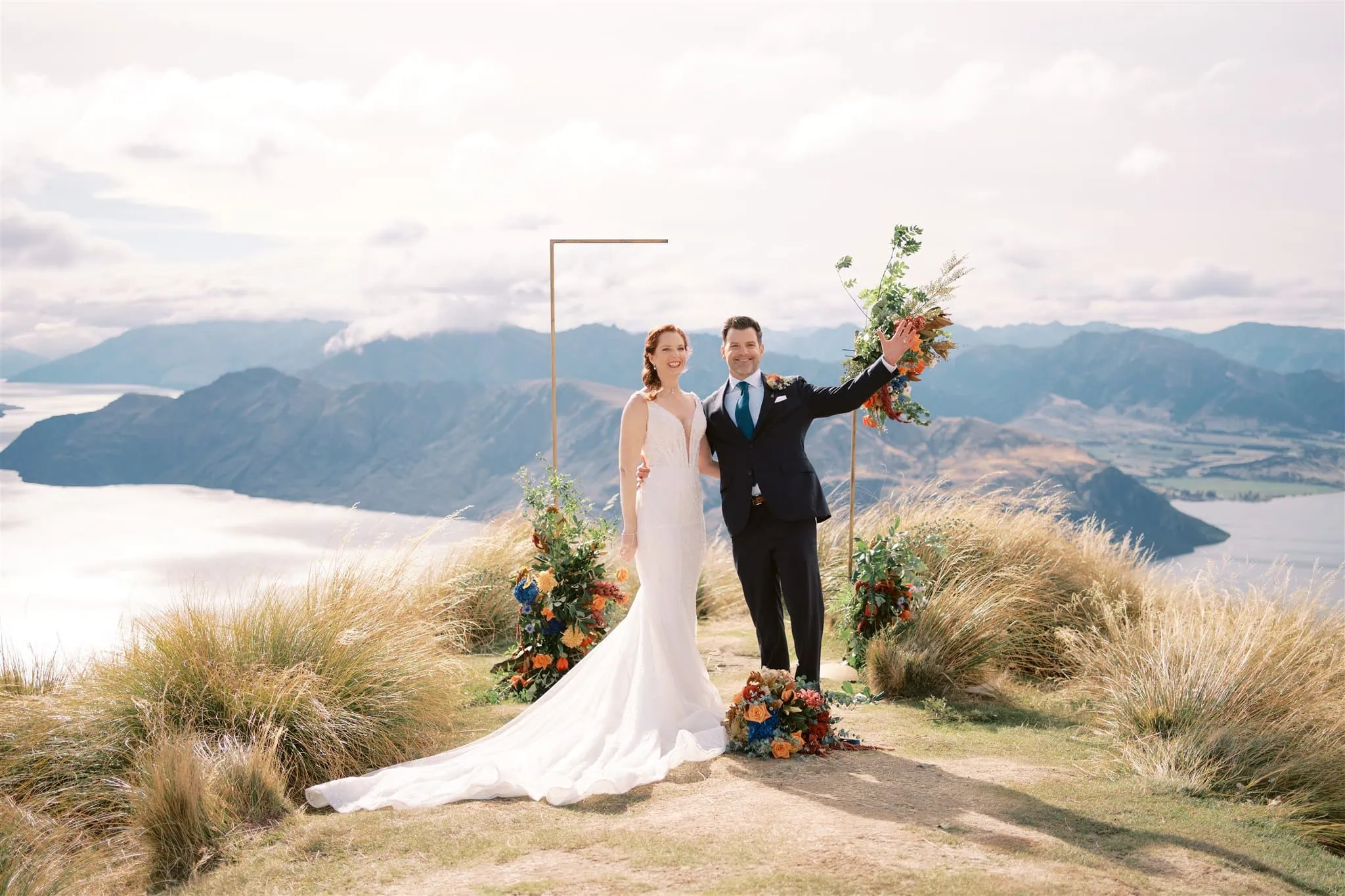 queenstown new zealand heli wedding elopement photographer videographer | A couple in wedding attire stands on a scenic hilltop with mountains and water in the background, capturing the essence of Queenstown elopement. They are smiling and waving, surrounded by beautiful floral arrangements—a perfect moment for wedding photography.