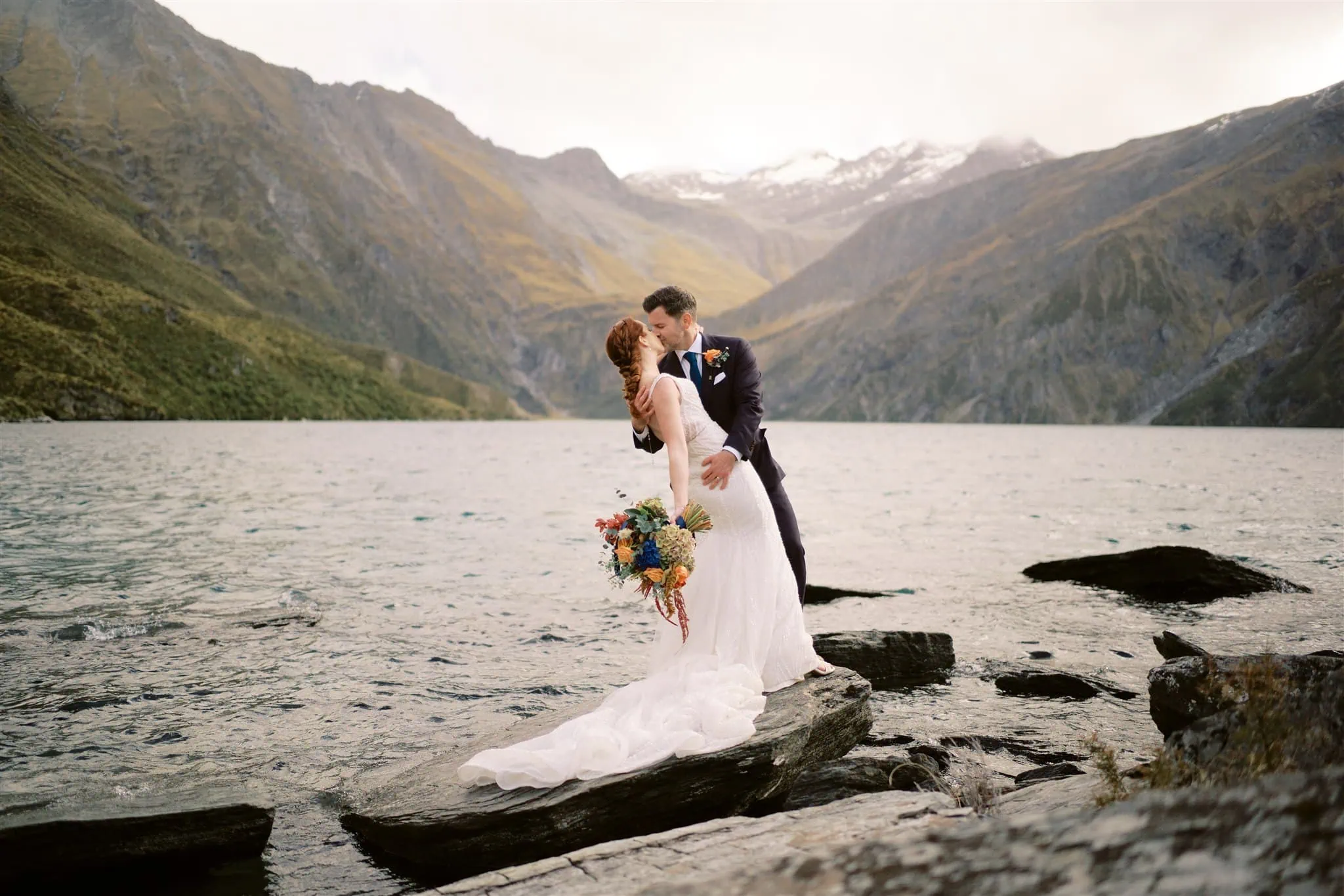 queenstown new zealand heli wedding elopement photographer videographer | A couple in wedding attire embraces on rocks by a lake, surrounded by mountains. The bride holds a bouquet, and the groom leans in for a kiss. This magical Queenstown elopement captures stunning wedding photography amidst nature's grandeur.