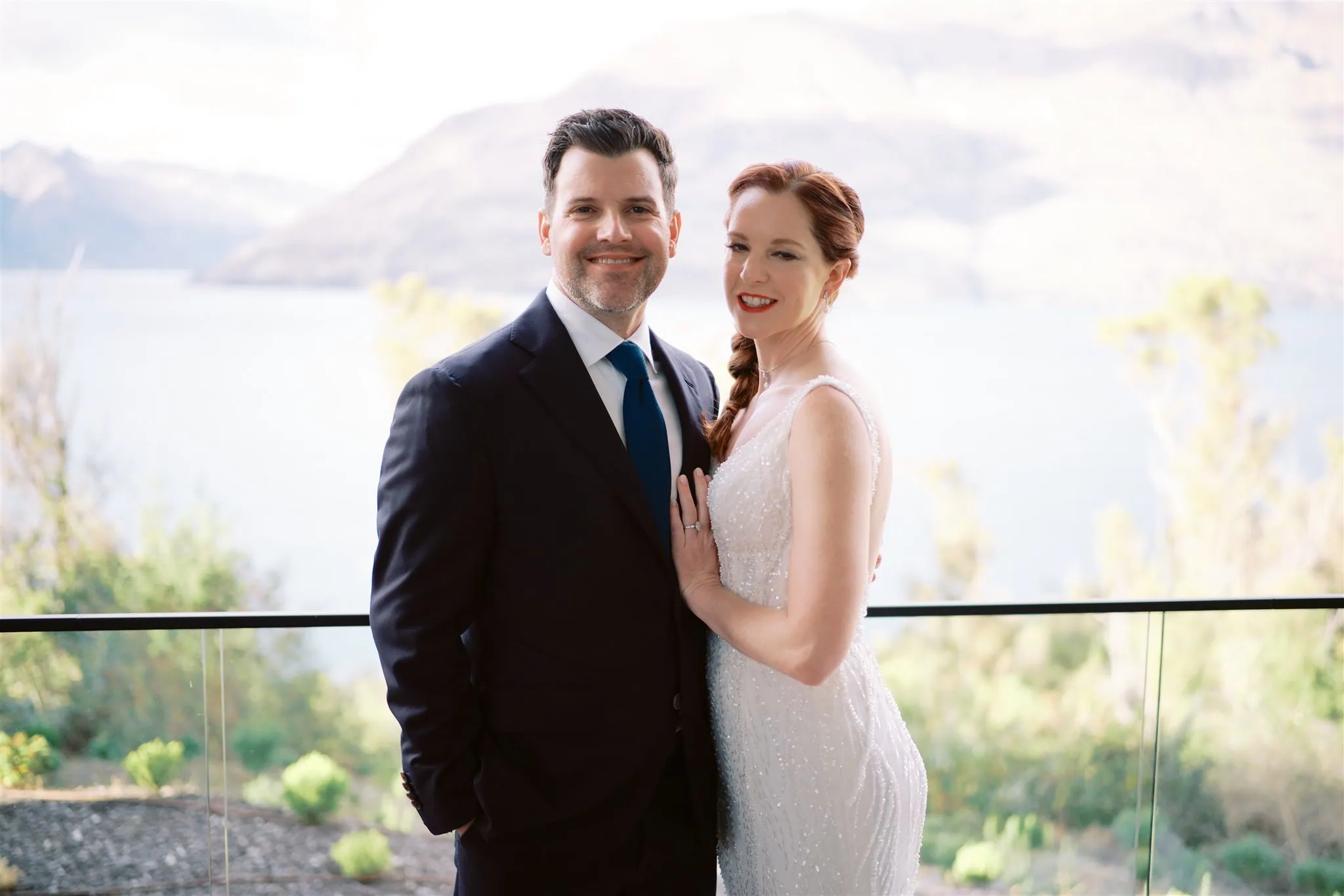 queenstown new zealand heli wedding elopement photographer videographer | A man in a suit and a woman in a white dress pose together on a balcony with a scenic backdrop of mountains and a lake, capturing the perfect moment of their Queenstown elopement wedding.