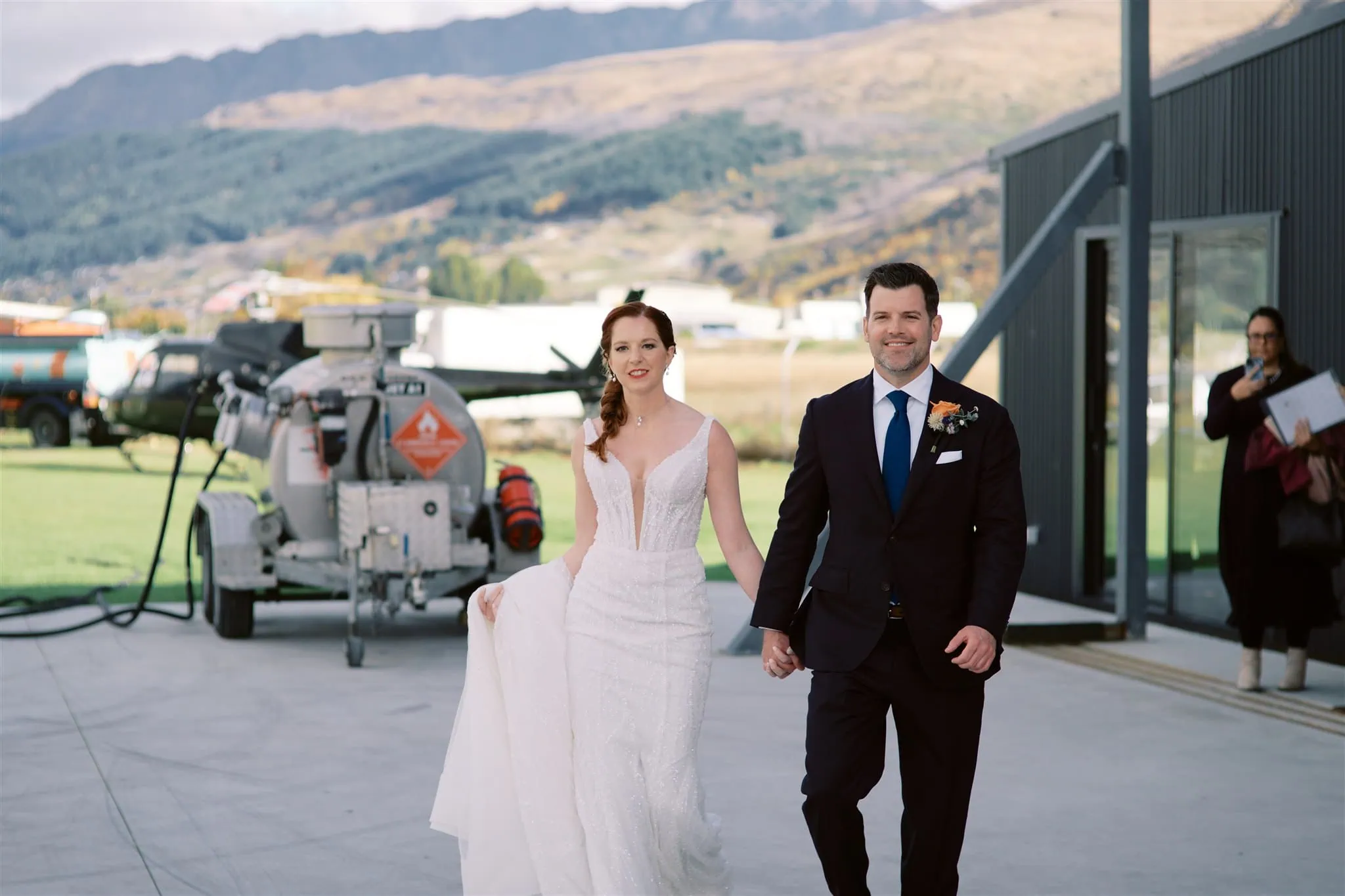 queenstown new zealand heli wedding elopement photographer videographer | A bride and groom holding hands walk outdoors near an aircraft hangar with mountains in the background, capturing the essence of a Queenstown Elopement. The bride wears a white dress and the groom dons a dark suit with a blue tie, making it perfect for Wedding Photography.