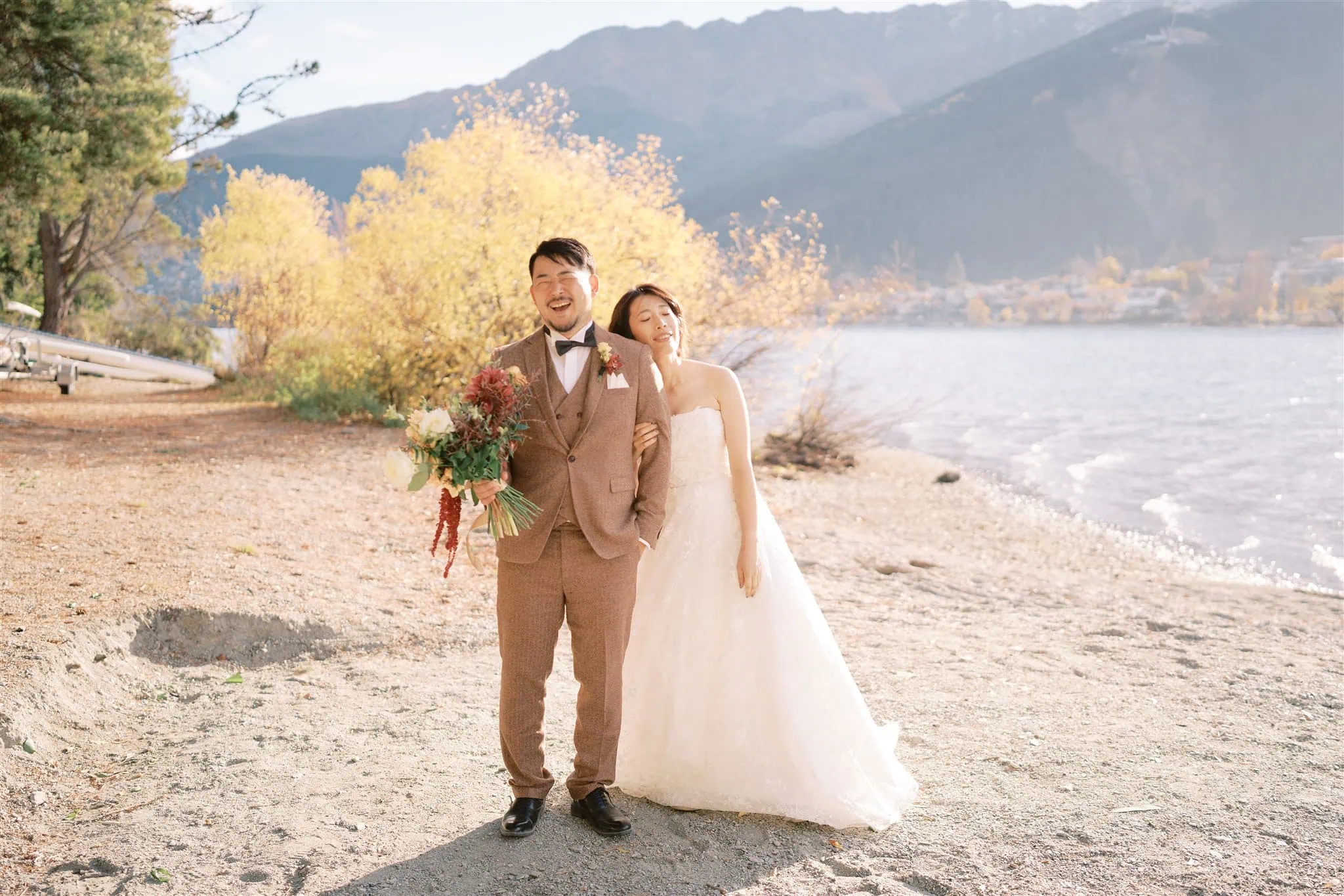 queenstown new zealand heli wedding elopement photographer videographer | A couple in formal attire stands on a lakeside beach in Queenstown. The man holds a bouquet, and they are both smiling. Trees with yellow leaves and mountains create a picturesque backdrop, perfect for an elopement wedding photography session.