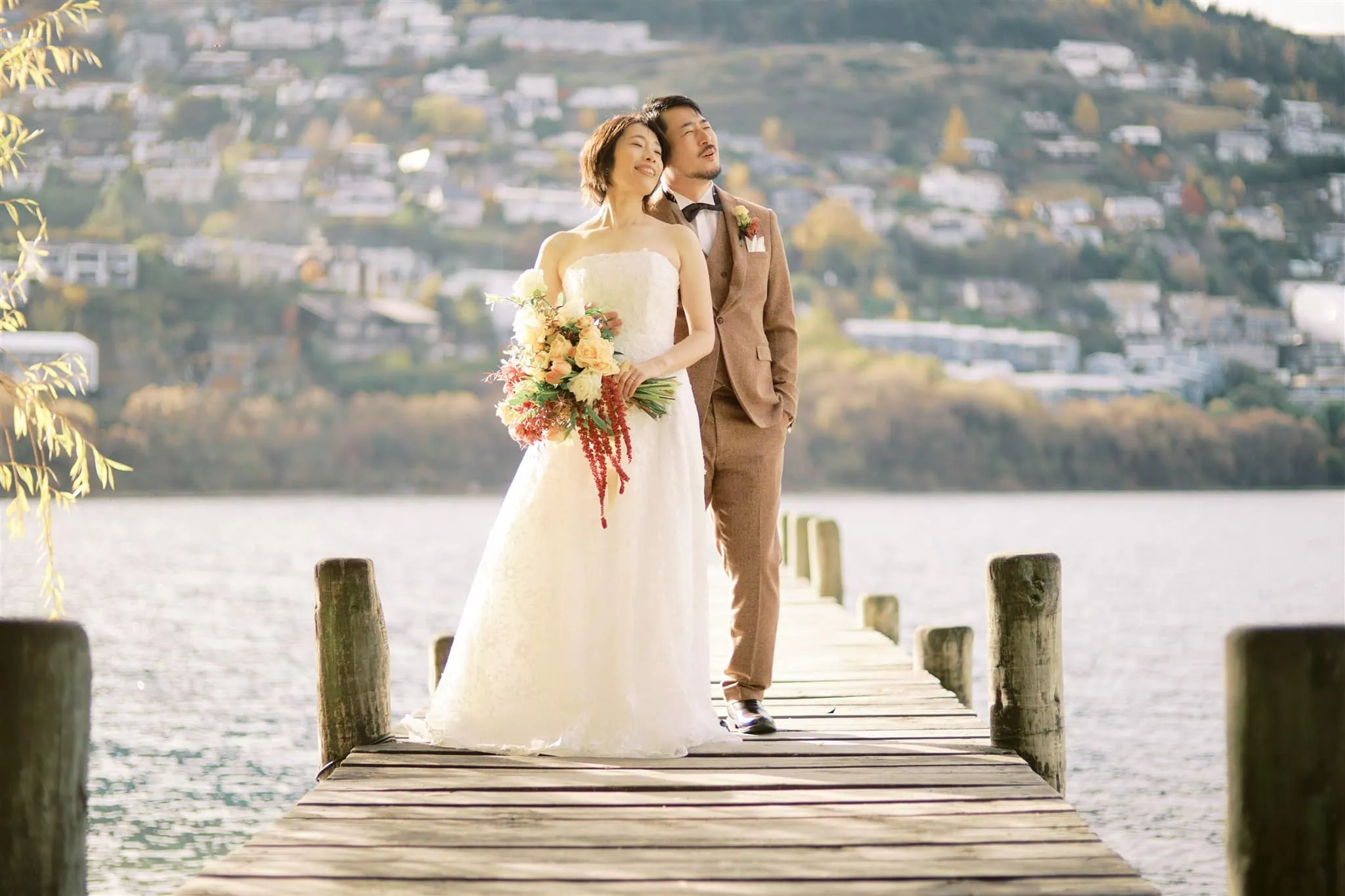 queenstown new zealand heli wedding elopement photographer videographer | A bride and groom stand on a wooden dock by a lake, dressed in a wedding gown and suit, looking into the distance with a forest and residential area in the background—a perfect scene for timeless wedding photography during their Queenstown elopement.