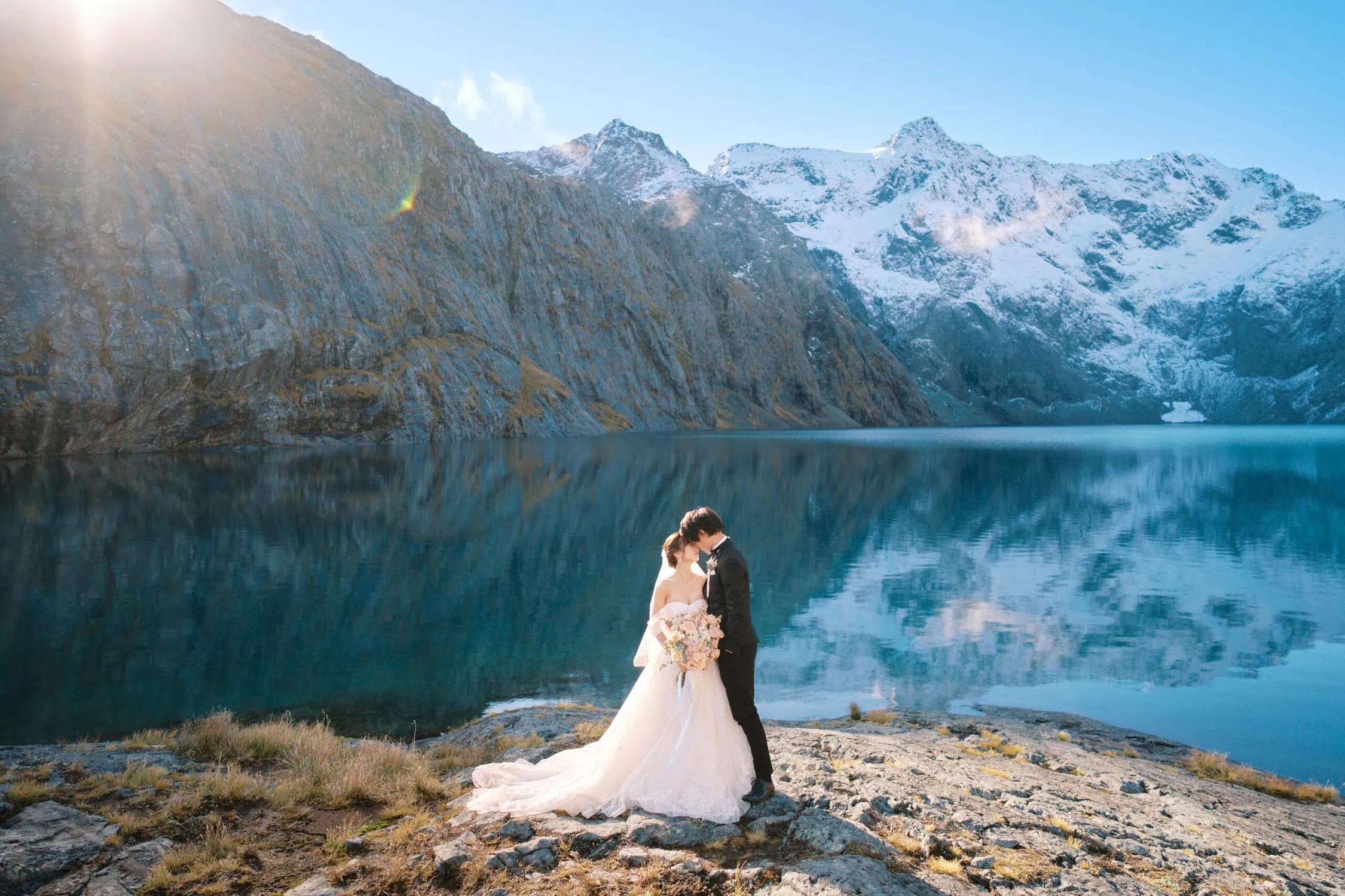 queenstown new zealand heli wedding elopement photographer videographer | A bride and groom stand embracing on a rocky shore, with a clear blue lake and snow-capped mountains in the background under a bright sky, capturing the essence of their Queenstown elopement.