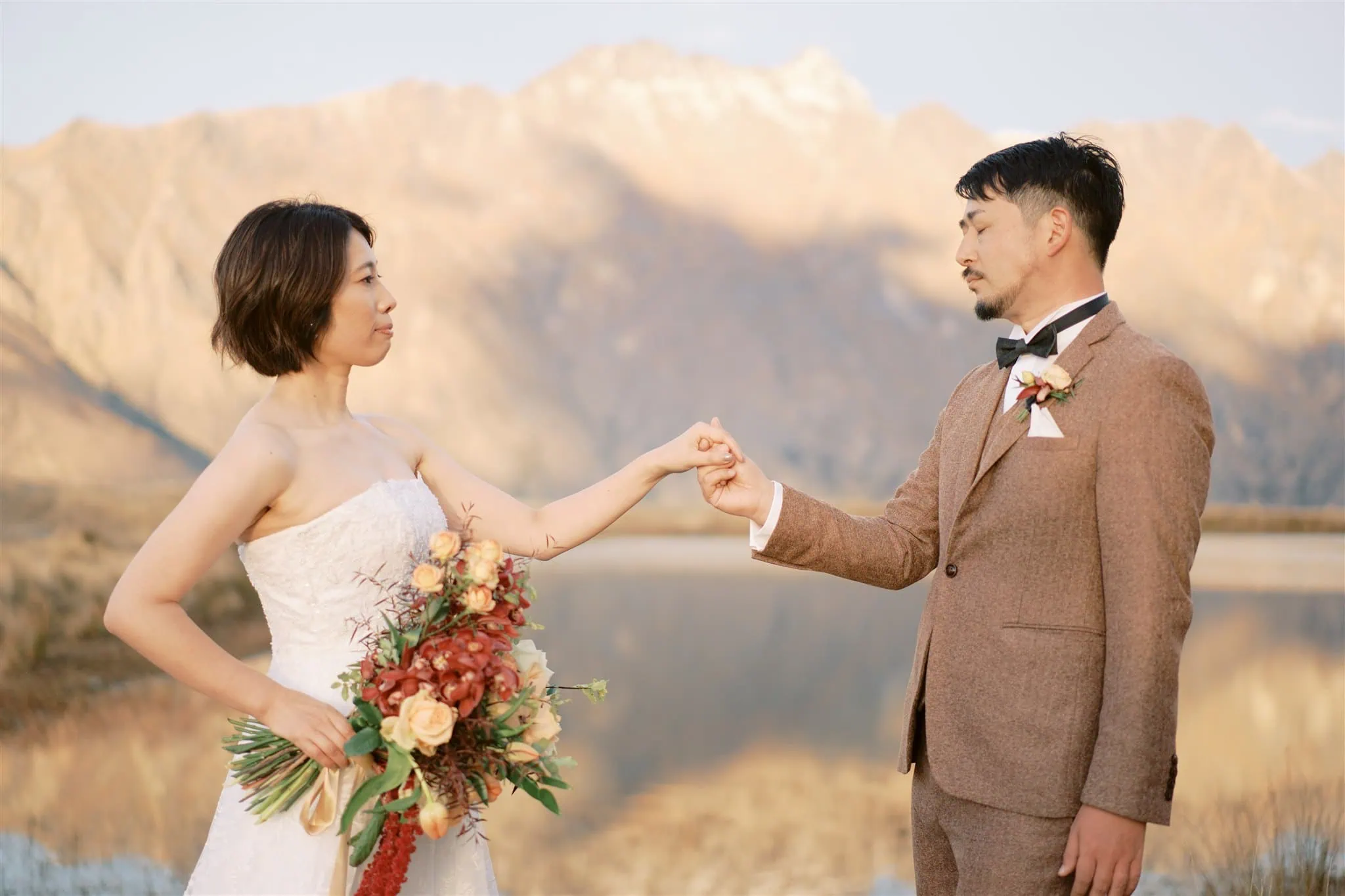 queenstown new zealand heli wedding elopement photographer videographer | A bride and groom dressed in a wedding gown and suit, respectively, stand in front of a serene pond in Queenstown with mountains in the background, touching their fists together. The bride holds a bouquet of flowers, capturing the essence of elopement wedding photography.