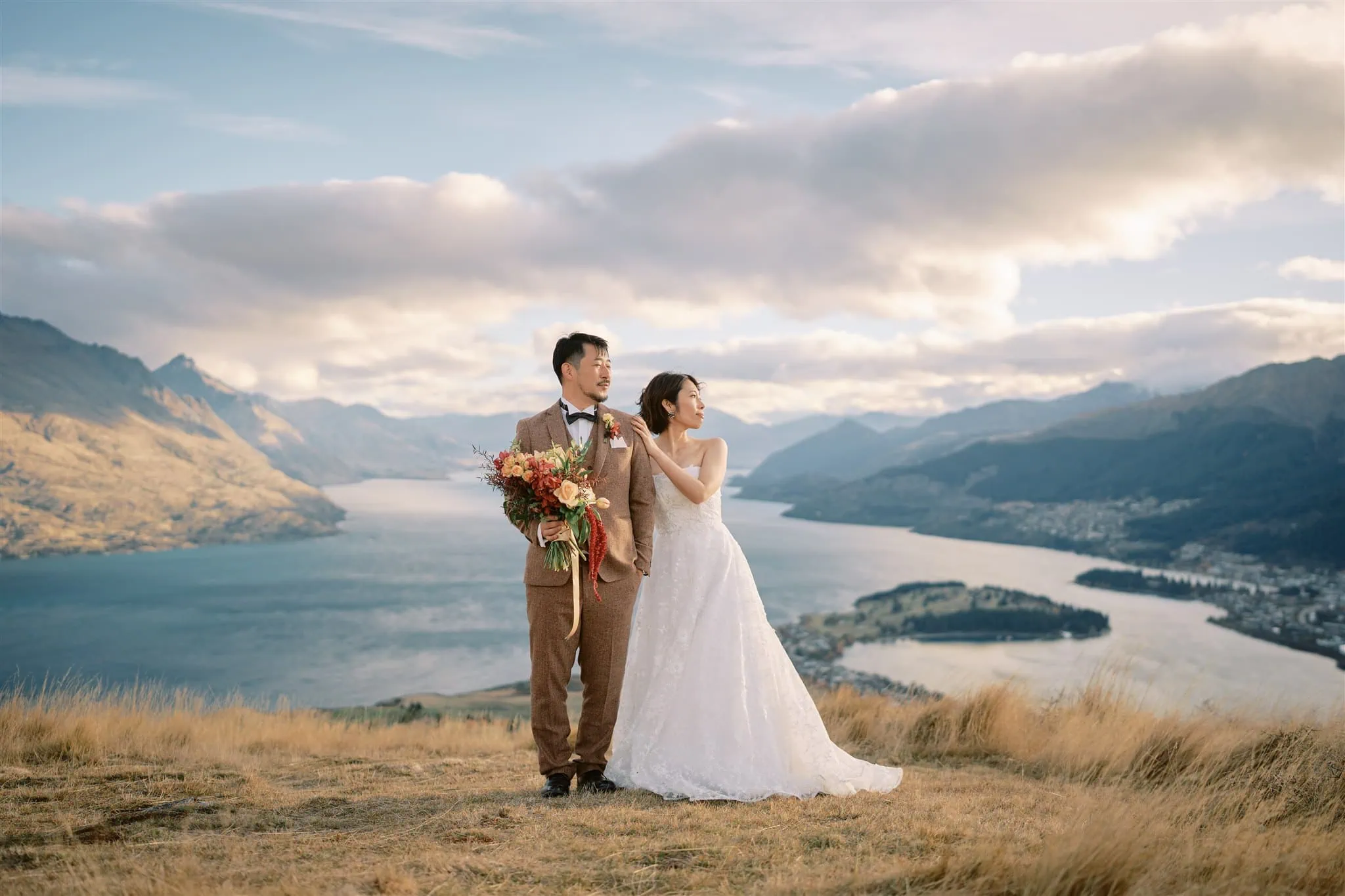 queenstown new zealand heli wedding elopement photographer videographer | A couple in wedding attire stands on a grassy hilltop with a scenic backdrop of a lake, mountains, and a partly cloudy sky during their Queenstown elopement. The bride holds a floral bouquet, capturing a perfect moment for wedding photography.