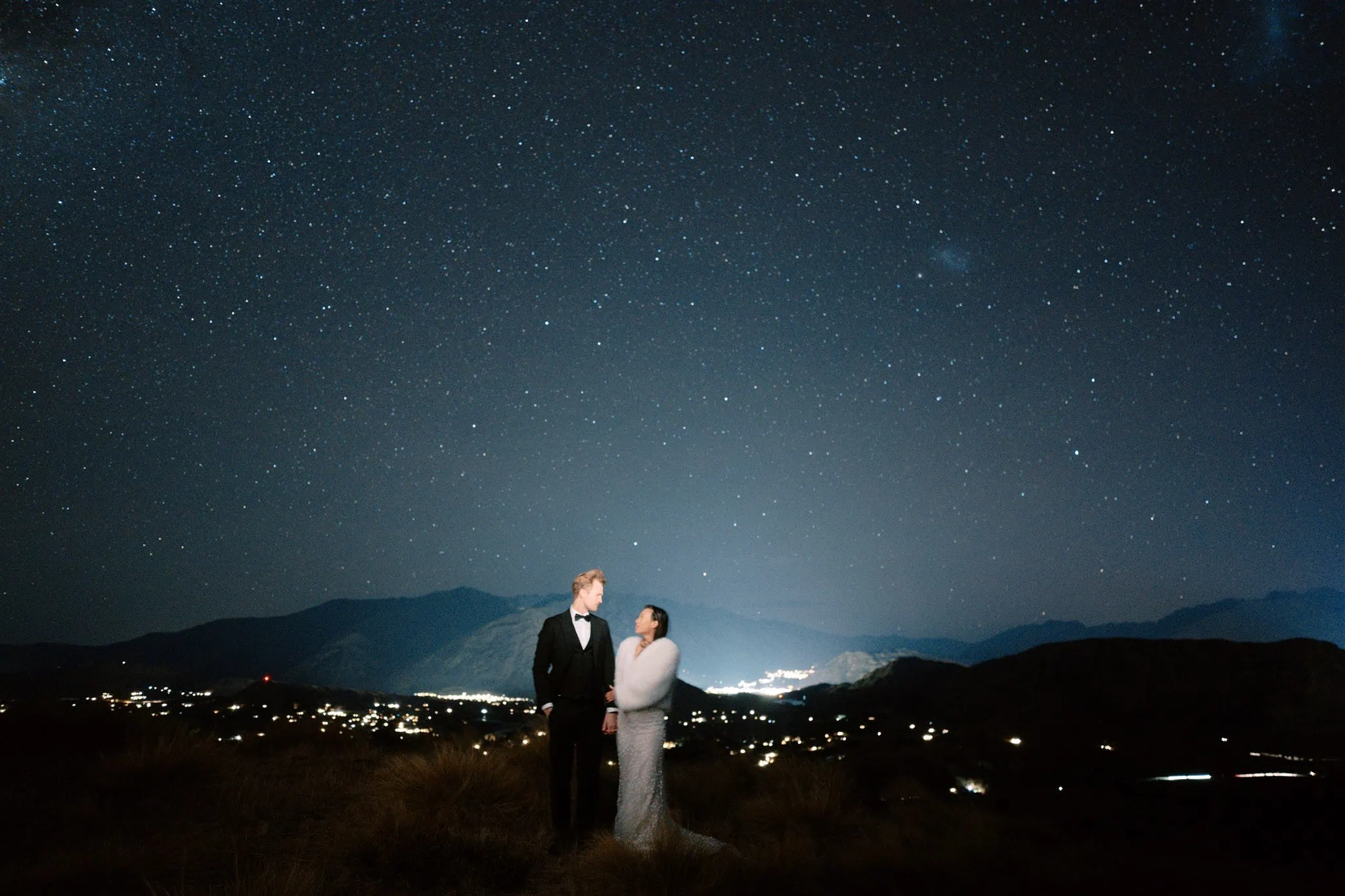 queenstown new zealand heli wedding elopement photographer videographer | A formally dressed couple stands together under a starry night sky with a cityscape in the background, capturing the magic of their Queenstown elopement through stunning wedding photography.