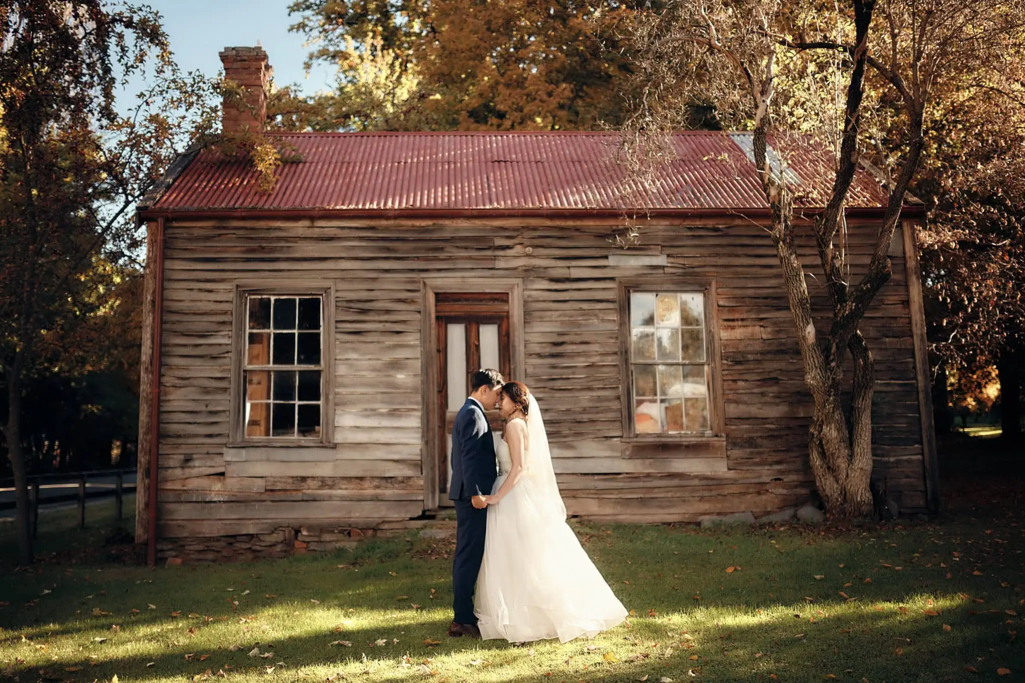 queenstown new zealand heli wedding elopement photographer videographer | A couple in wedding attire stands close together on a lawn, in front of an old, weathered wooden house with a rusty metal roof and trees surrounding it—a perfect scene for Nicholas & Tingyi's pre-wedding photography.