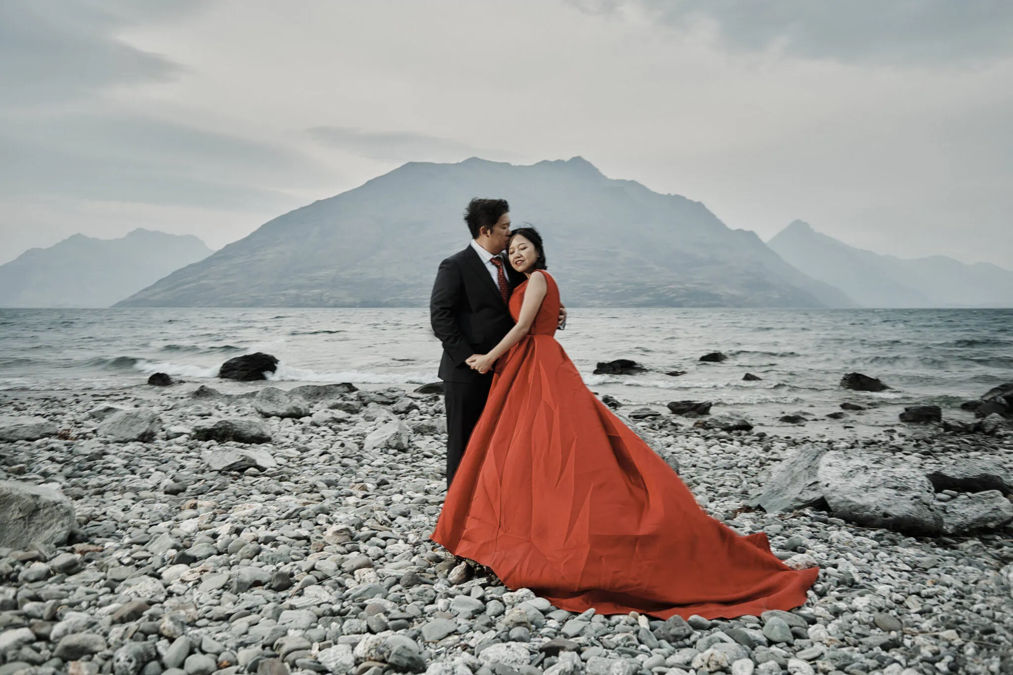 queenstown new zealand heli wedding elopement photographer videographer | Jun Yang & Ida, dressed in formal attire with Ida in a stunning red gown, pose on a rocky shore for their pre-wedding photography session. With Queenstown's majestic mountains and serene waters as their backdrop, the scene captures their love beautifully.