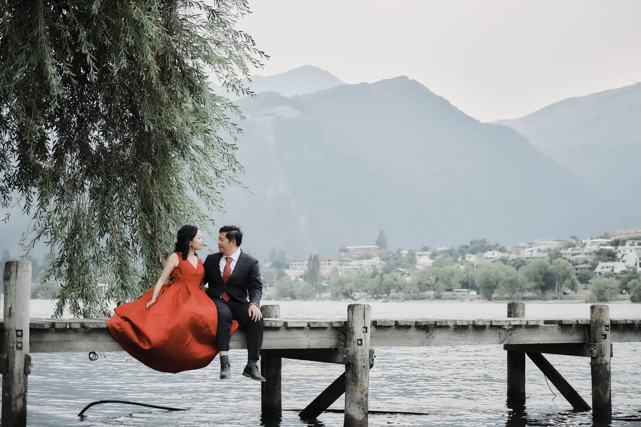 queenstown new zealand heli wedding elopement photographer videographer | A couple in formal attire sits on a wooden pier over a lake with a mountainous landscape in the background, captured beautifully by Jun Yang. The woman, Ida, wears a red gown, and the man wears a suit. This memorable moment forever immortalized as part of their Queenstown Pre-wedding Photography.