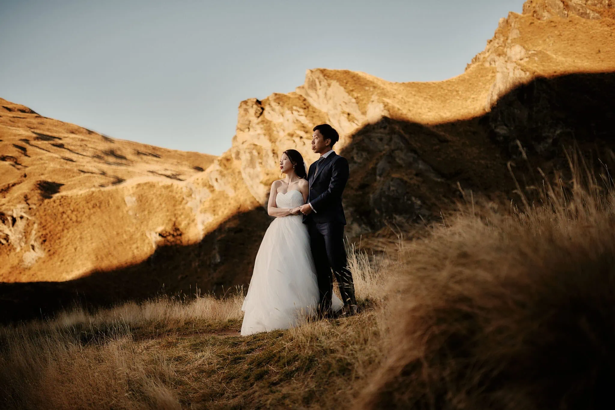 queenstown new zealand heli wedding elopement photographer videographer | Nicholas & Tingyi stand together, gazing into the distance in a mountainous landscape under clear skies on a sunlit dirt path with dry grass and rocky hills in the background, beautifully captured for their Queenstown pre-wedding photography.