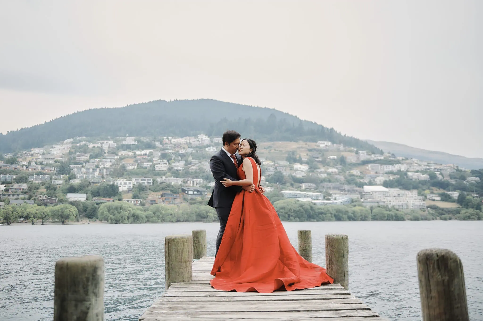 queenstown new zealand heli wedding elopement photographer videographer | A couple stands on a wooden dock by a lake, with the man in a dark suit and the woman in a red gown. A town and hills can be seen in the background. Captured beautifully by Jun Yang, this Queenstown pre-wedding shoot encapsulates love amidst stunning scenery.