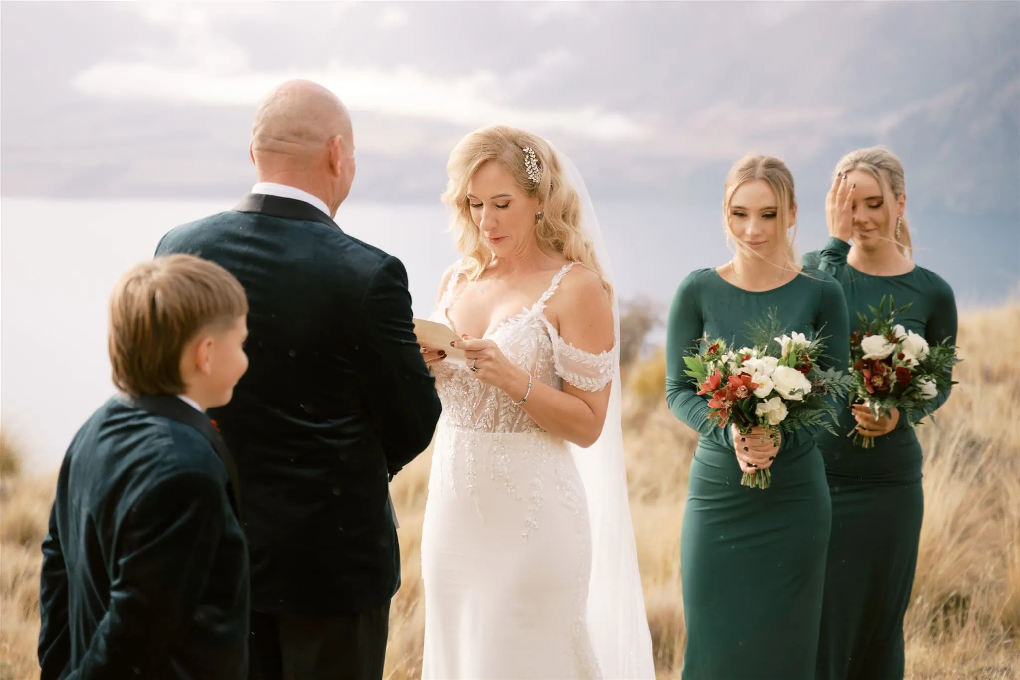 queenstown new zealand heli wedding elopement photographer videographer | At a breathtaking Queenstown wedding, a bride reads her vows to the groom during an intimate outdoor ceremony. Captured by expert wedding photography, two bridesmaids clutch vibrant bouquets while a young boy stands nearby, witnessing their love.