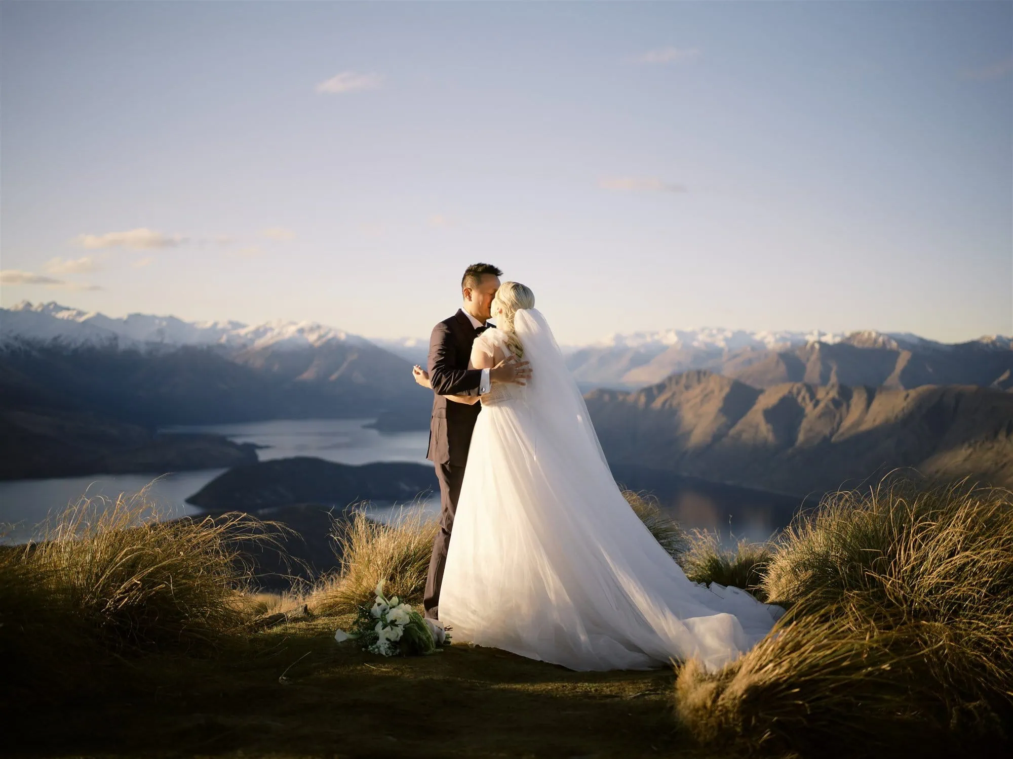 queenstown new zealand heli wedding elopement photographer videographer | Georgina & Ageng share a tender kiss on a mountaintop, capturing the essence of elopement wedding photography. The breathtaking backdrop of Queenstown's serene lake and majestic mountains under a clear sky makes their moment truly unforgettable.
