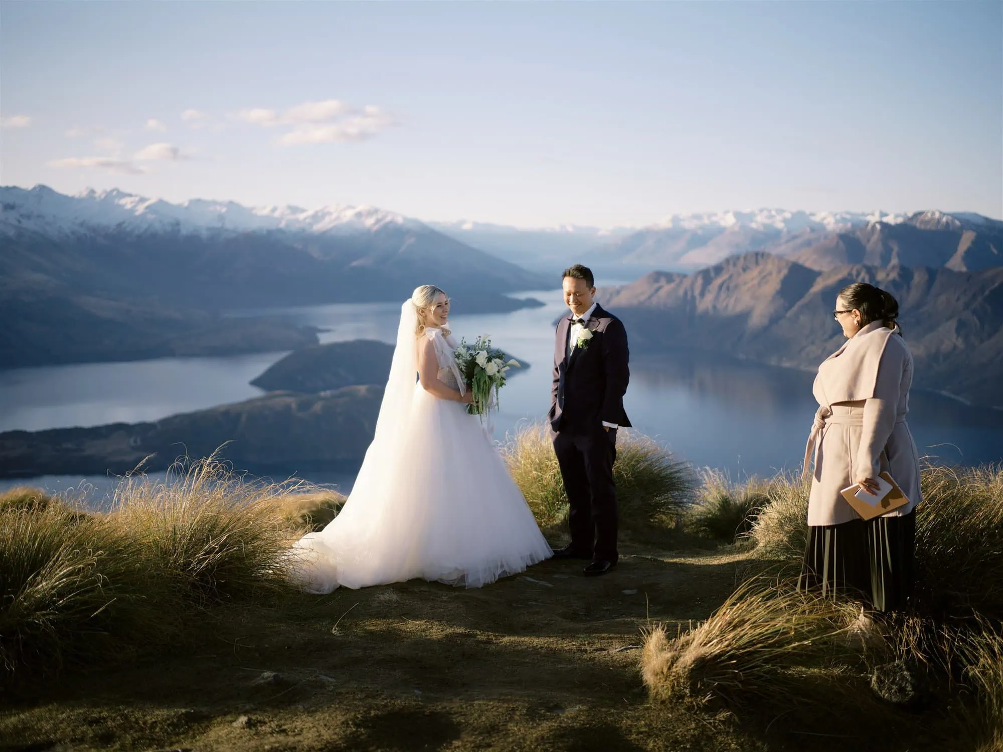 queenstown new zealand heli wedding elopement photographer videographer | A couple is eloping on a grassy hilltop in Queenstown, with a stunning lake and mountain backdrop. The bride holds a bouquet, the groom stands beside her, while a wedding photographer captures every moment as the officiant holds a book nearby.