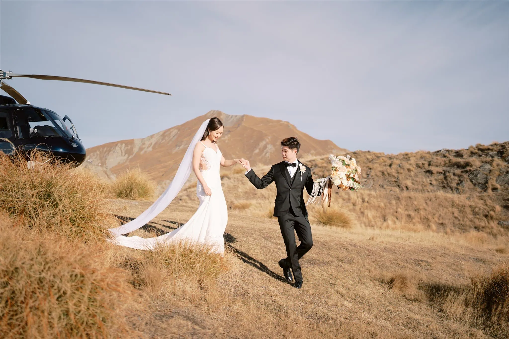 queenstown new zealand heli wedding elopement photographer videographer | A bride and groom walk hand in hand on a grassy hillside during their elopement wedding, with the majestic mountains of Queenstown and a helicopter in the background, perfectly capturing the essence of their special day in stunning photography.