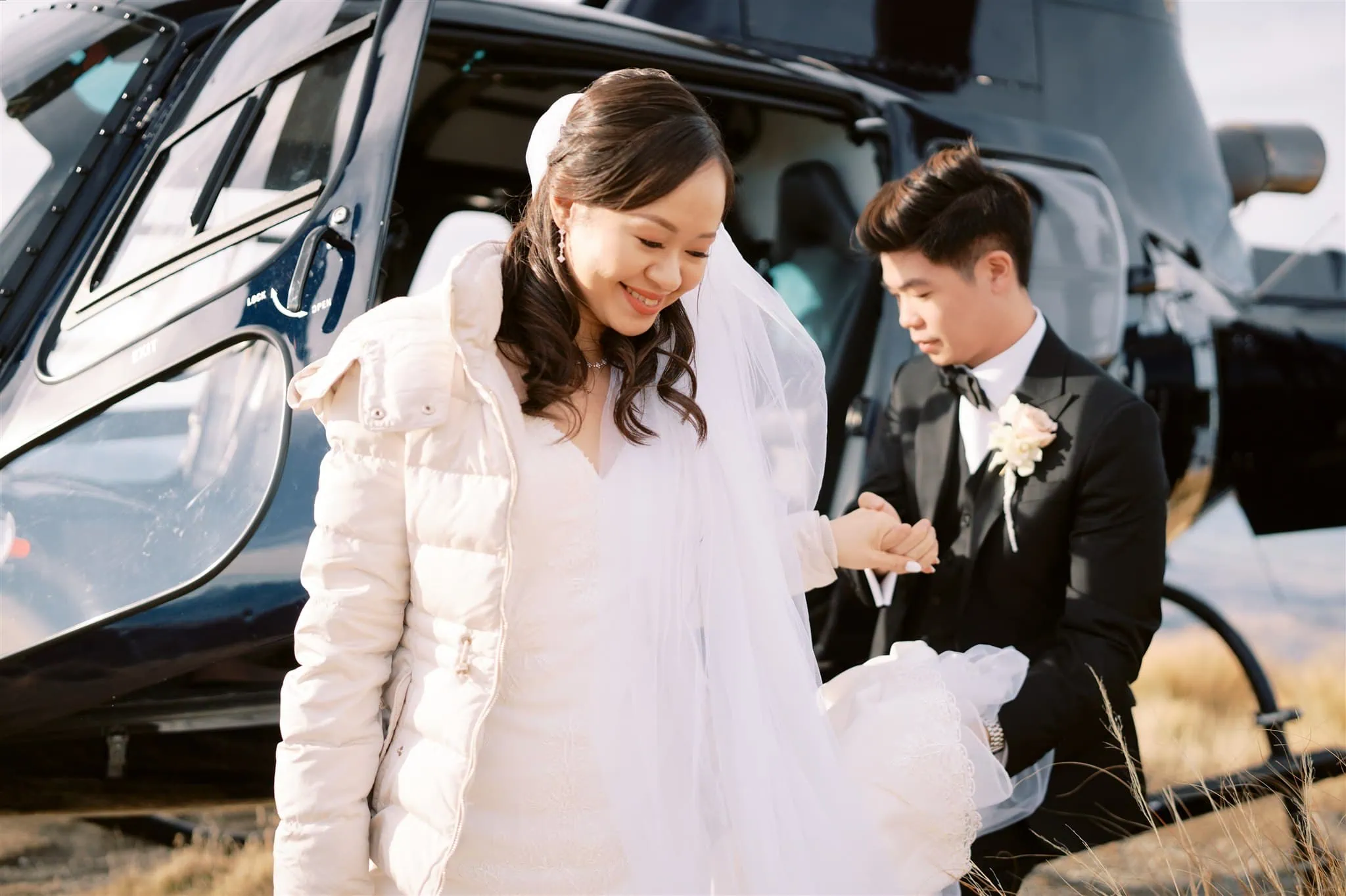 queenstown new zealand heli wedding elopement photographer videographer | A smiling bride in a white dress and coat steps out of a helicopter, assisted by a groom in a black suit and bow tie, set against the stunning backdrop of Queenstown—a perfect moment captured for an elopement wedding photography session.
