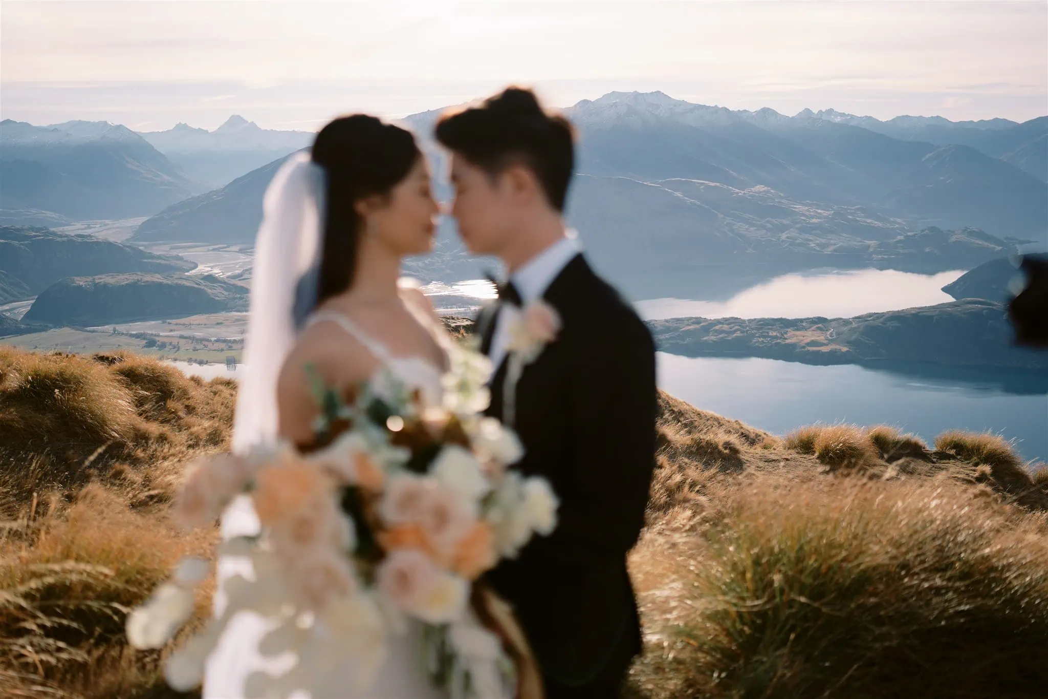 queenstown new zealand heli wedding elopement photographer videographer | Tiffany and Malcolm stand close together, holding a bouquet, with a scenic mountain and lake view in the background, capturing their Queenstown elopement through stunning wedding photography.