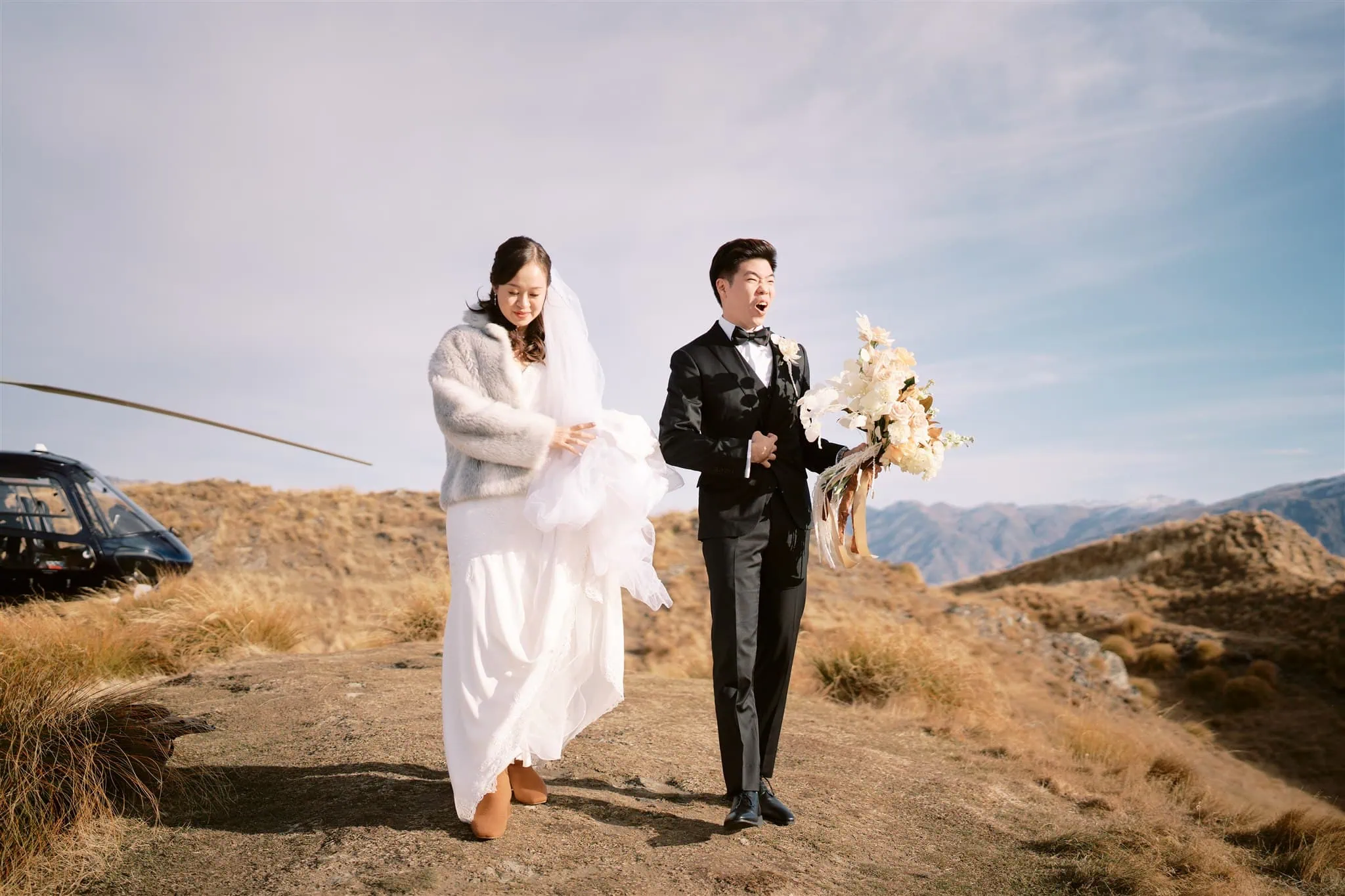 queenstown new zealand heli wedding elopement photographer videographer | A bride and groom walk along a rocky path with a helicopter in the backdrop, framed by Queenstown’s grassy hills under a blue sky. The groom clutches a bouquet, capturing the essence of intimate elopement and breathtaking wedding photography.
