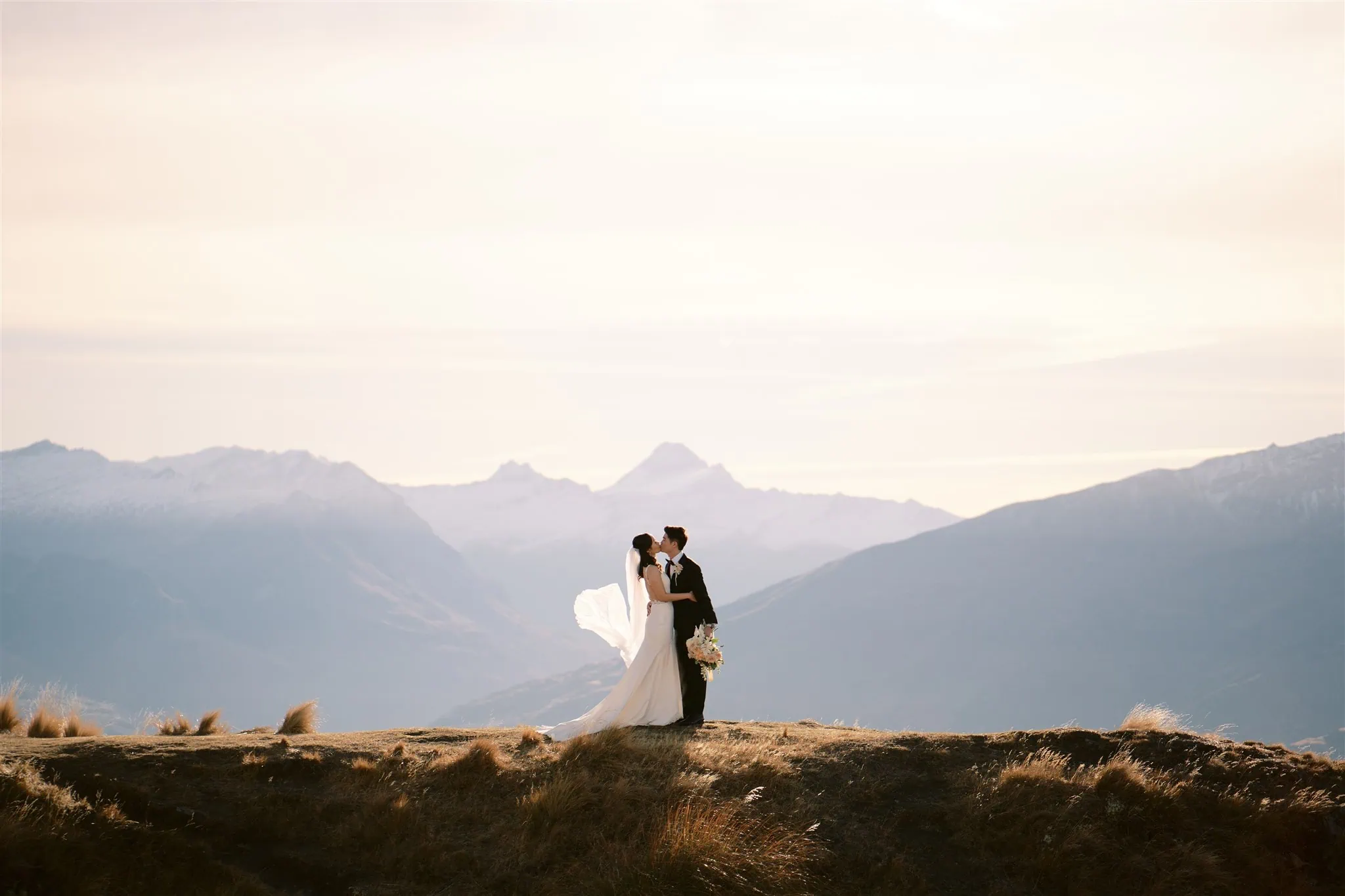 queenstown new zealand heli wedding elopement photographer videographer | A bride and groom share a kiss on a mountain ridge in Queenstown, with distant peaks enhancing the moment against the clear sky—a perfect setting for stunning wedding photography.