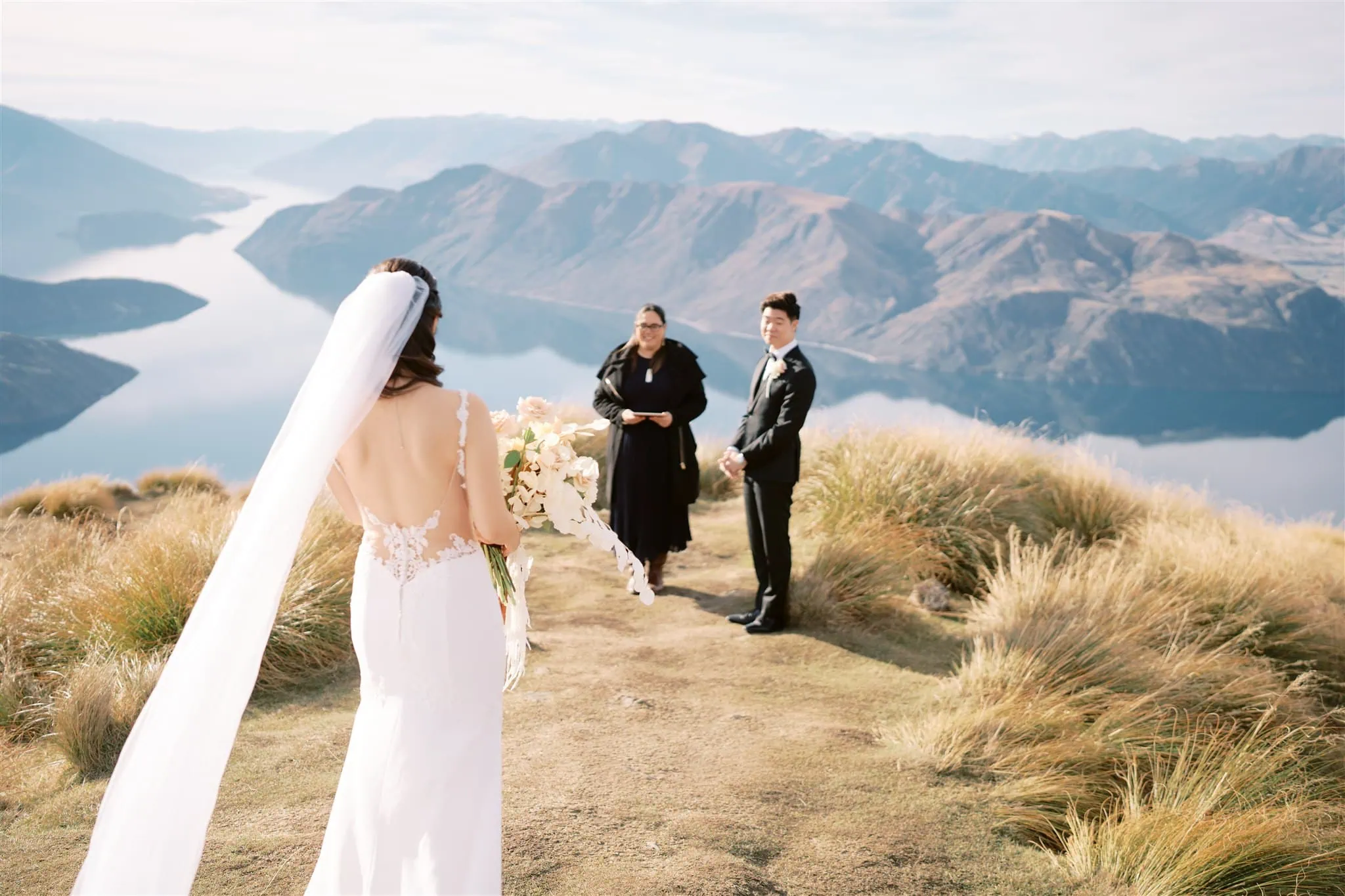 queenstown new zealand heli wedding elopement photographer videographer | A Bride and groom, Tiffany & Malcolm, stand with an officiant on a grassy hilltop overlooking a lake and mountains in Queenstown Elopement style, ready for their wedding ceremony. The bride holds a bouquet amidst the stunning scenery waiting for the Wedding Photography to capture perfect moments.
