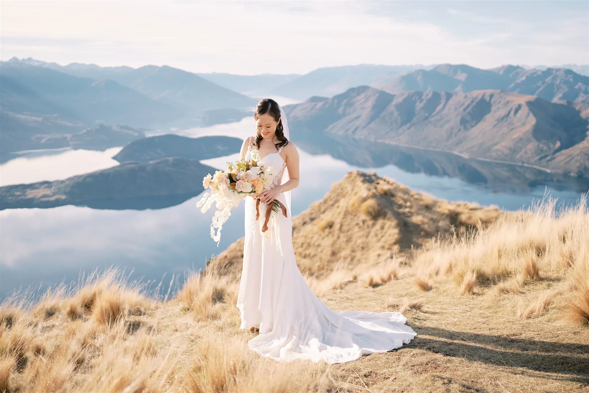 queenstown new zealand heli wedding elopement photographer videographer | Bride in a white gown holding a bouquet stands on a grassy hilltop with a scenic lake and mountain view in the background, capturing the magic of Tiffany & Malcolm's Queenstown elopement.
