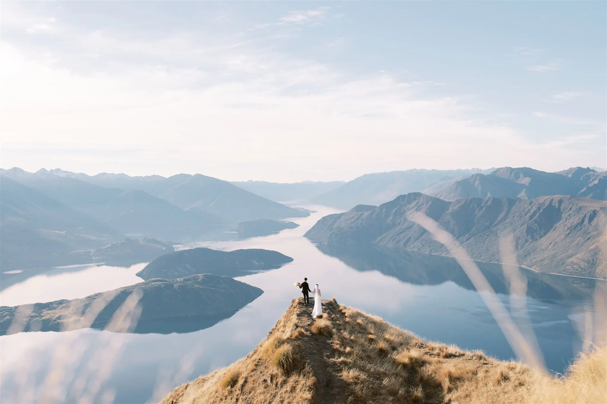 queenstown new zealand heli wedding elopement photographer videographer | Tiffany & Malcolm stand hand in hand on a grassy hill, overlooking the vast lake and majestic mountains of their Queenstown elopement. The clear sky above serves as the perfect backdrop for their unforgettable wedding photography.