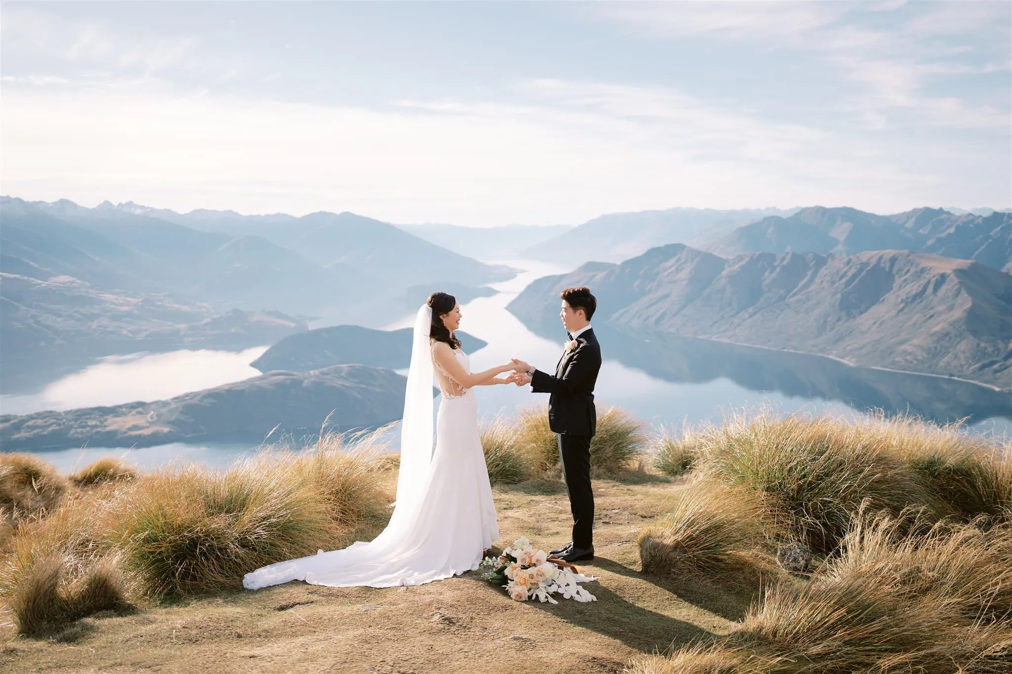 queenstown new zealand heli wedding elopement photographer videographer | On a grassy hilltop in Queenstown, a bride and groom stand facing each other, embraced by the stunning backdrop of mountains and lake. Their moment, perfectly captured by photography, whispers the essence of an intimate elopement wedding. Nearby, a bouquet rests gently on the ground.