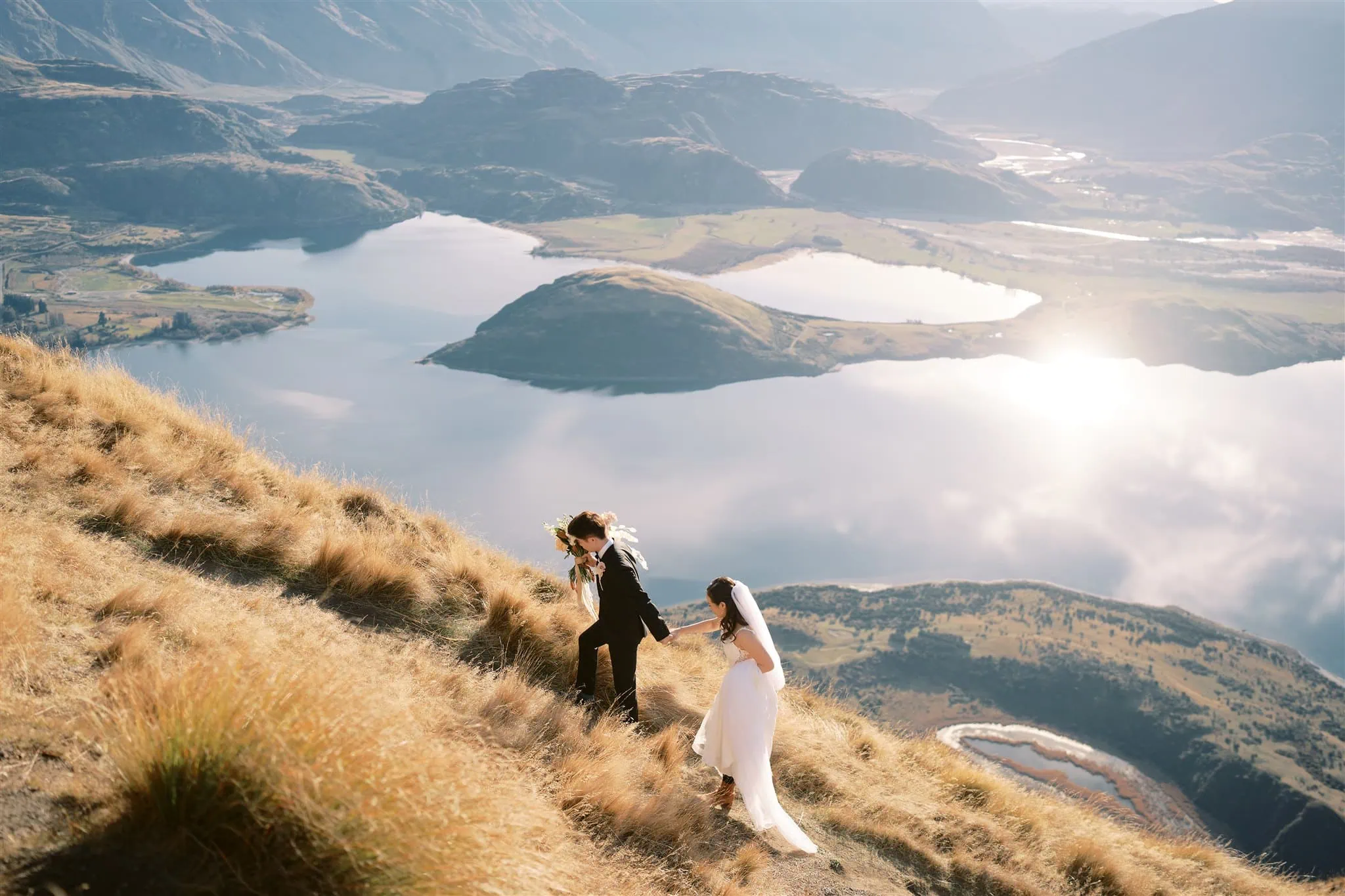queenstown new zealand heli wedding elopement photographer videographer | A couple in wedding attire ascends a grassy hill, capturing the essence of an elopement with the scenic beauty of Queenstown's lake and mountains in the background, perfect for timeless wedding photography.