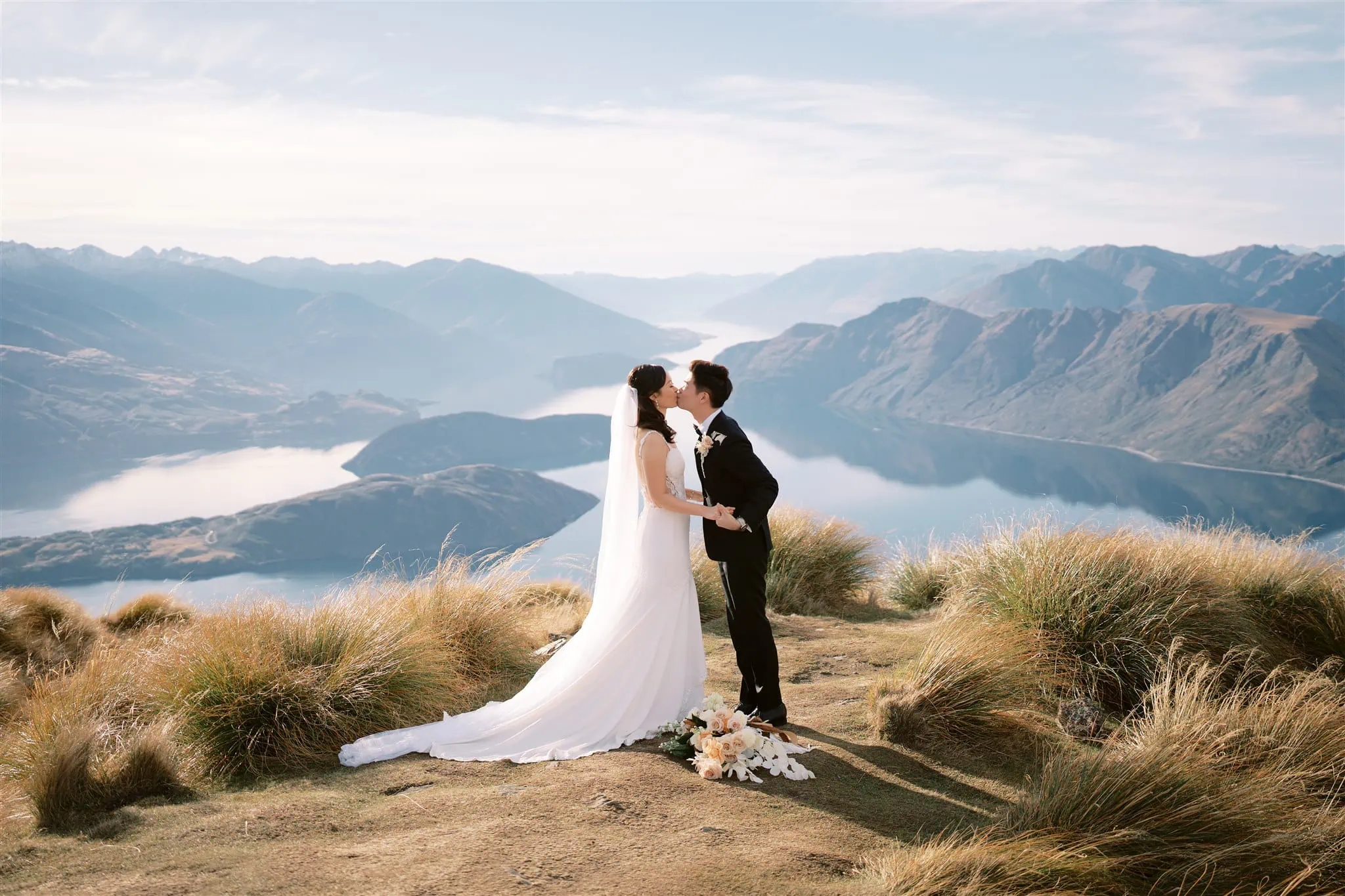 queenstown new zealand heli wedding elopement photographer videographer | A bride and groom share a kiss on a grassy hilltop in Queenstown, surrounded by scenic mountains and a shimmering lake, capturing the essence of perfect wedding photography.
