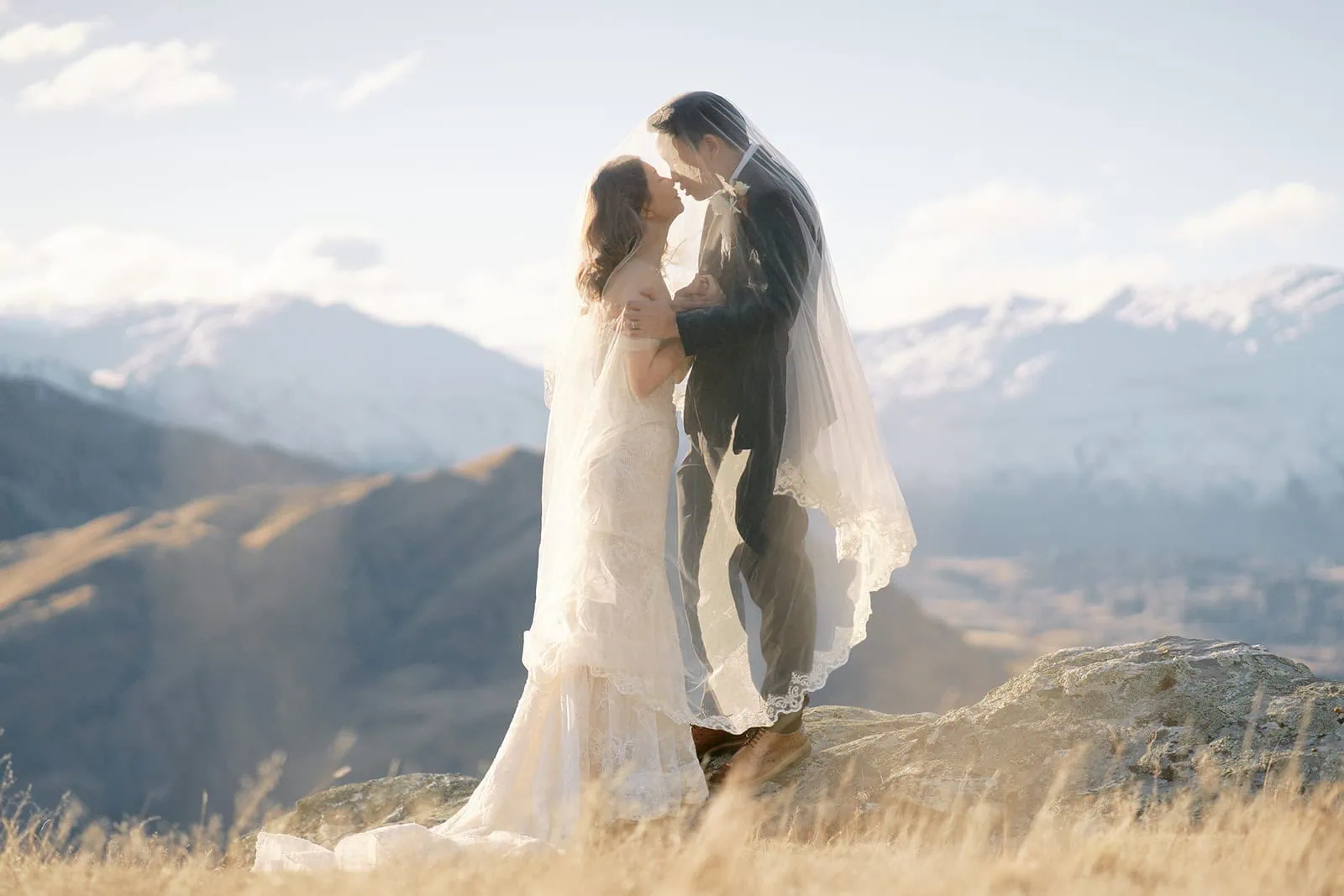 queenstown new zealand heli wedding elopement photographer videographer | Ryan and Crystal stand on a rocky ledge in Queenstown, embracing under a shared veil. The mountains in the background create the perfect setting for their enchanting pre-wedding photography moment.