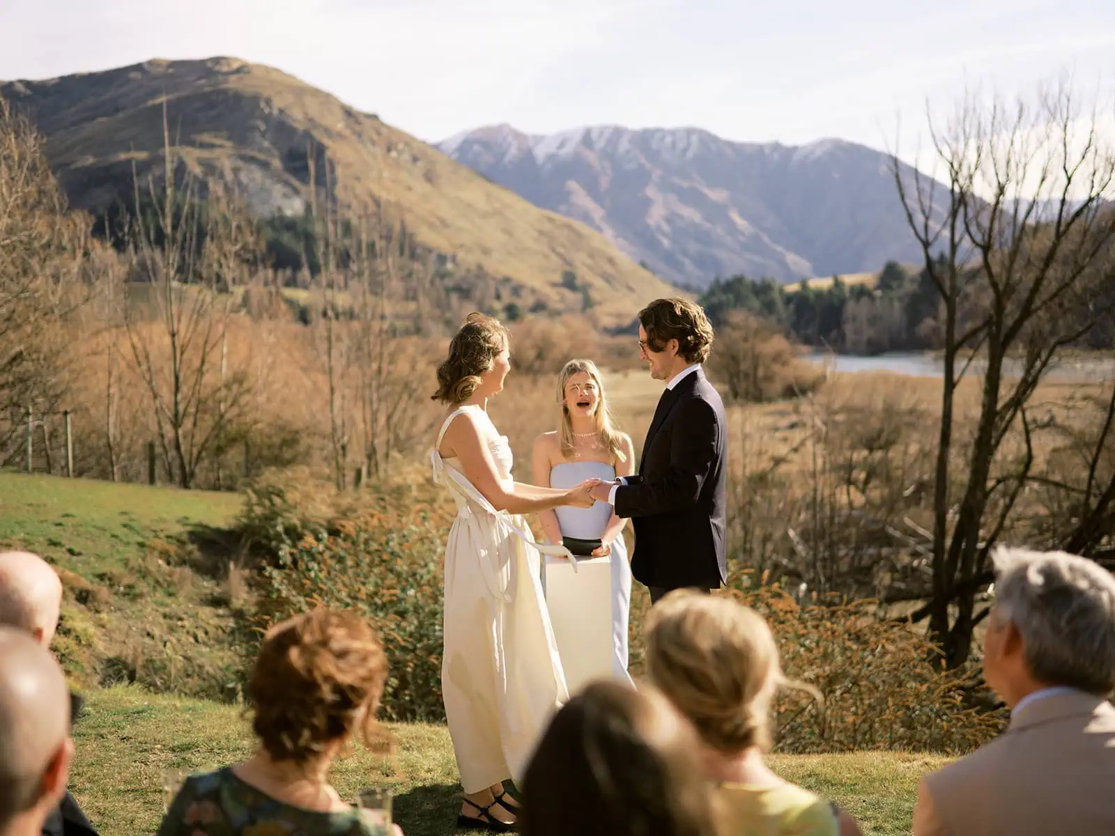 queenstown new zealand heli wedding elopement photographer videographer | In a serene Queenstown elopement, a couple exchanges vows outdoors with an officiant. Guests sit in the foreground as hills and trees frame the backdrop, capturing the essence of wedding photography perfection.