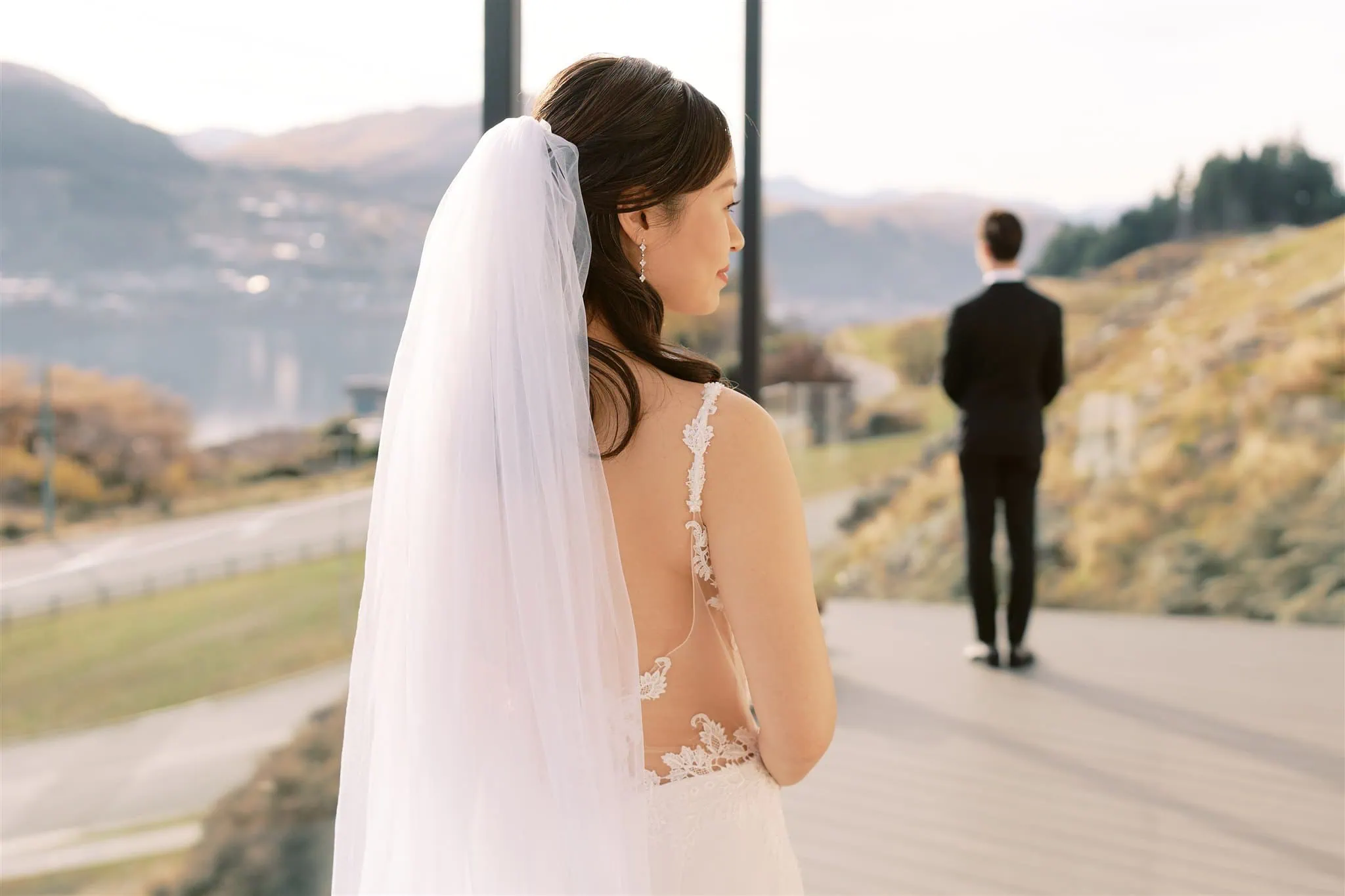 queenstown new zealand heli wedding elopement photographer videographer | A bride in a white dress and veil stands outdoors, gazing at Malcolm in a suit facing away, set against Queenstown's scenic backdrop—a perfect capture of their intimate elopement by wedding photography experts.