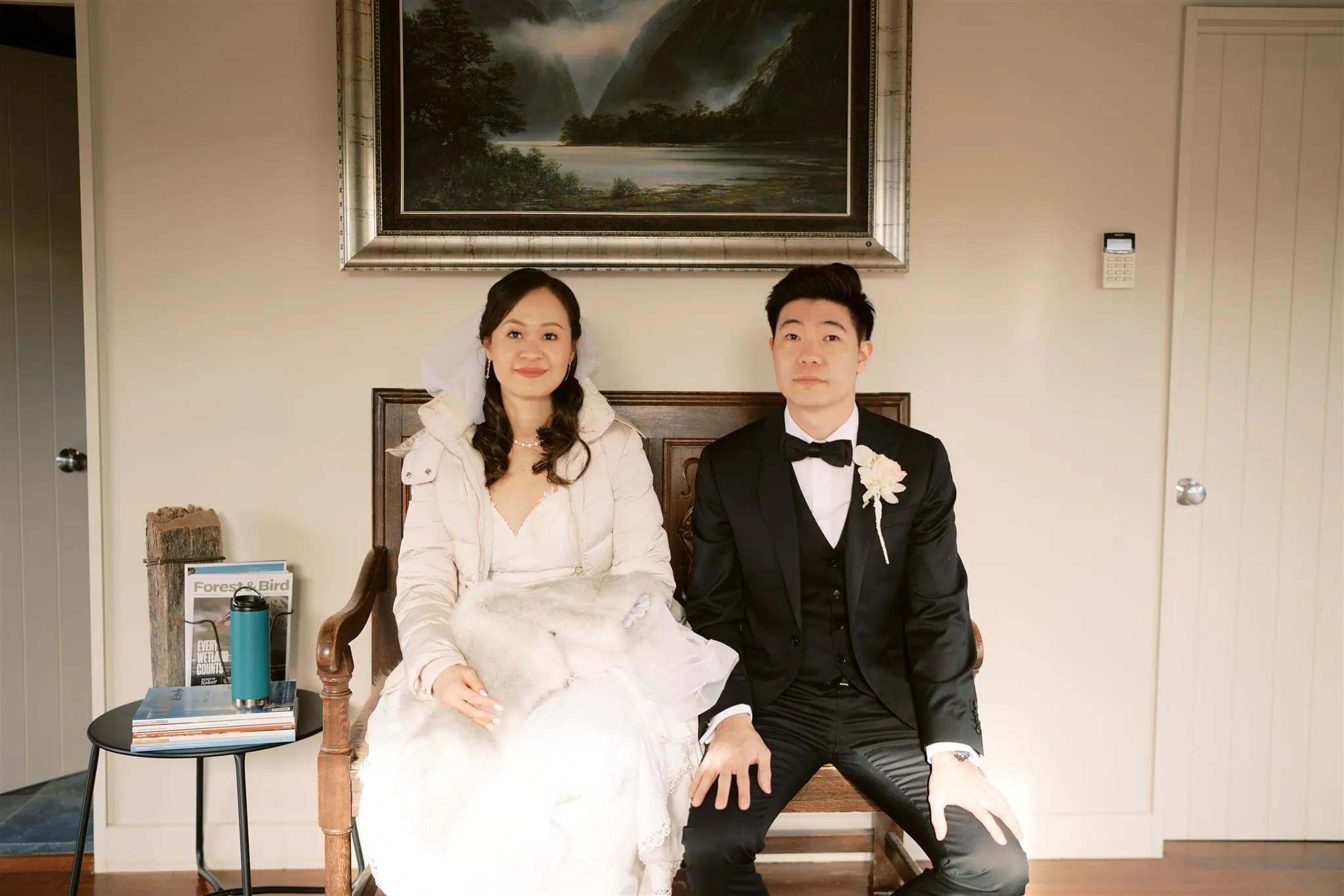 queenstown new zealand heli wedding elopement photographer videographer | A bride and groom sit on a bench in a room with a framed landscape painting on the wall, capturing the essence of Queenstown's beauty. The bride wears a veil and white dress, symbolizing their intimate elopement; the groom complements her in a black suit and bow tie—a perfect scene for wedding photography.