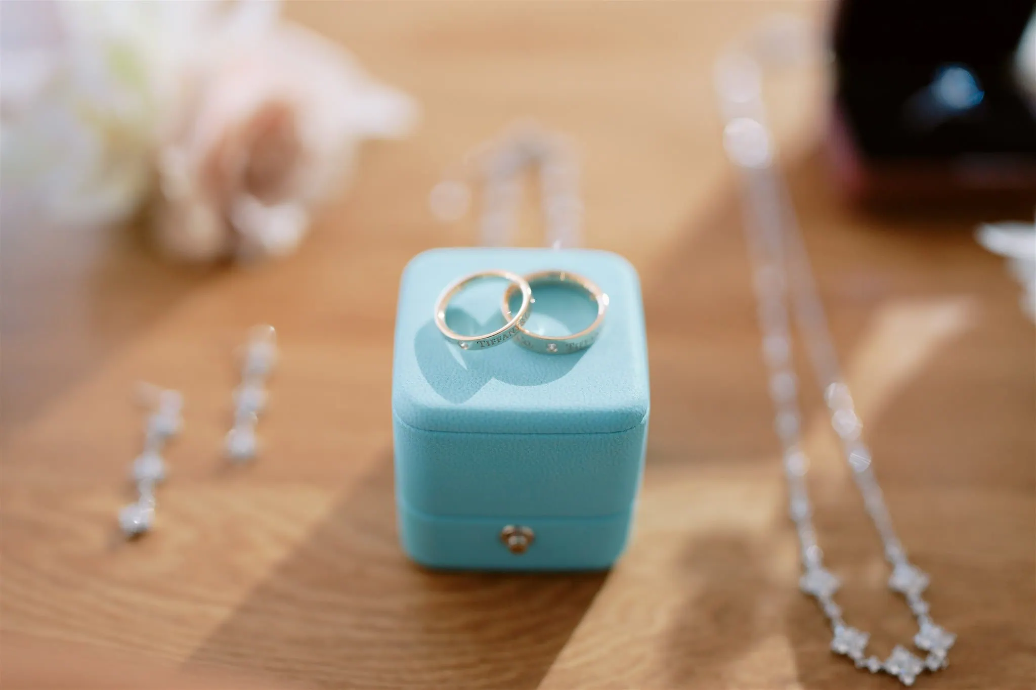 queenstown new zealand heli wedding elopement photographer videographer | Two gold rings on a turquoise jewelry box, captured beautifully by Queenstown Elopement Wedding Photography, are surrounded by earrings, a necklace, and a blurred flower in the background.