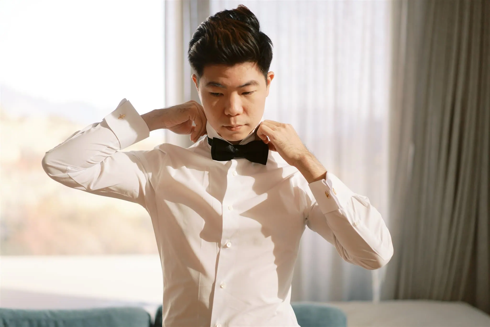 queenstown new zealand heli wedding elopement photographer videographer | In a bright room with elegant curtains, Tiffany adjusts Malcolm's black bow tie. This intimate moment is beautifully captured as part of their Queenstown elopement, showcasing the essence of wedding photography.