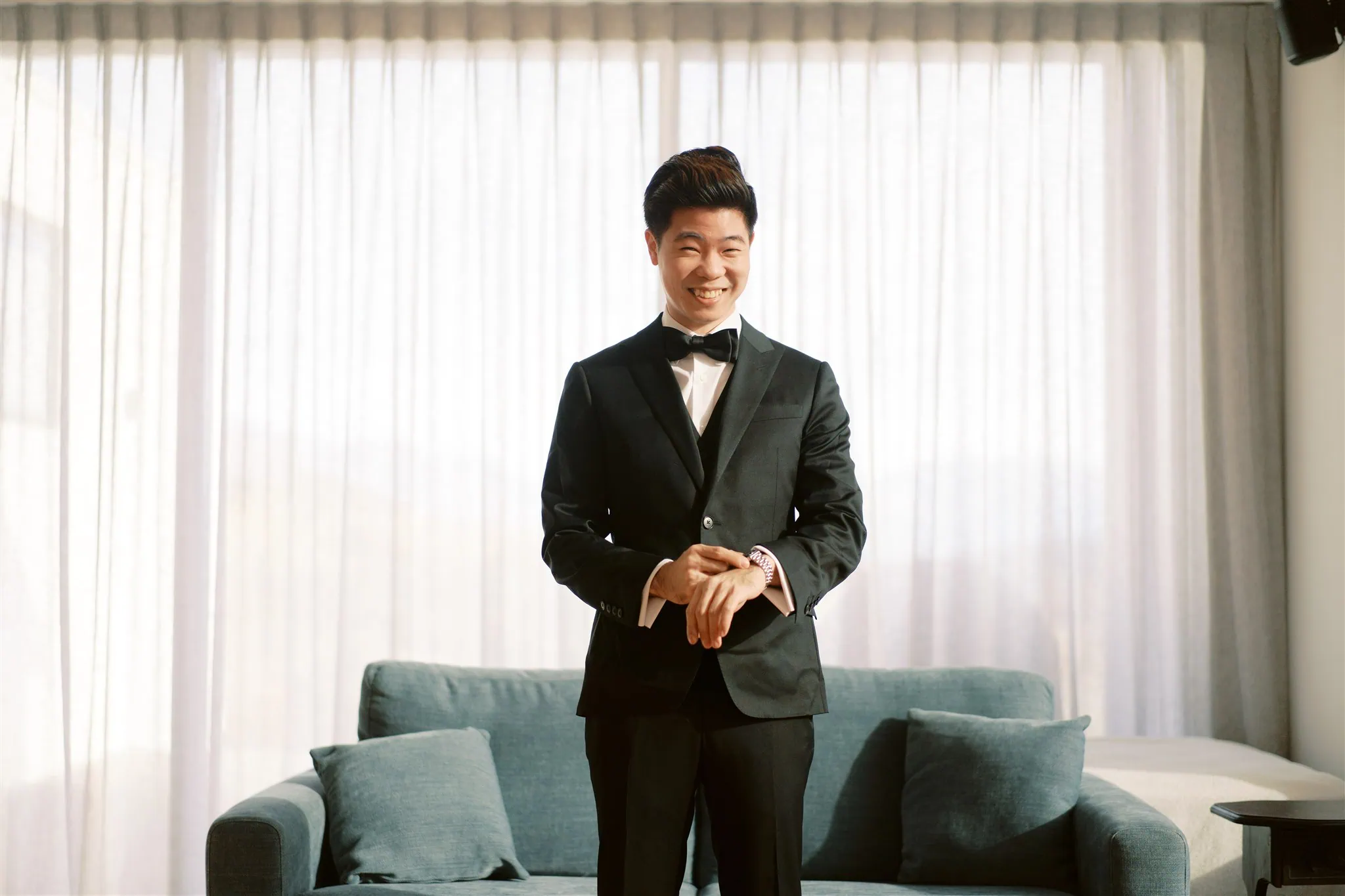 queenstown new zealand heli wedding elopement photographer videographer | A person in a black suit and bow tie stands smiling in front of a sofa and curtained window, capturing the elegant simplicity of an elopement wedding.