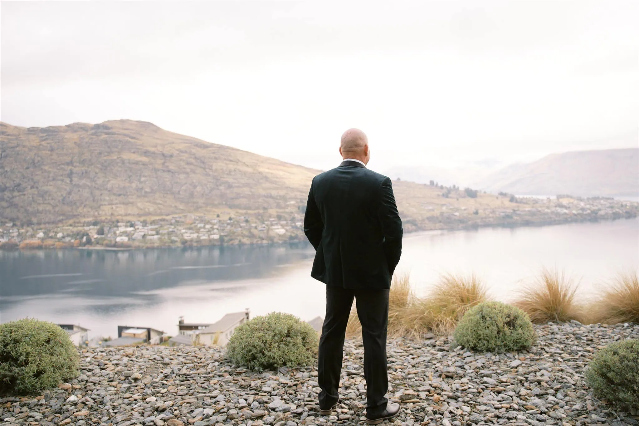 queenstown new zealand heli wedding elopement photographer videographer | A man in a suit stands on rocky terrain, overlooking a calm lake and hills under a cloudy sky, capturing the essence of a Queenstown elopement.