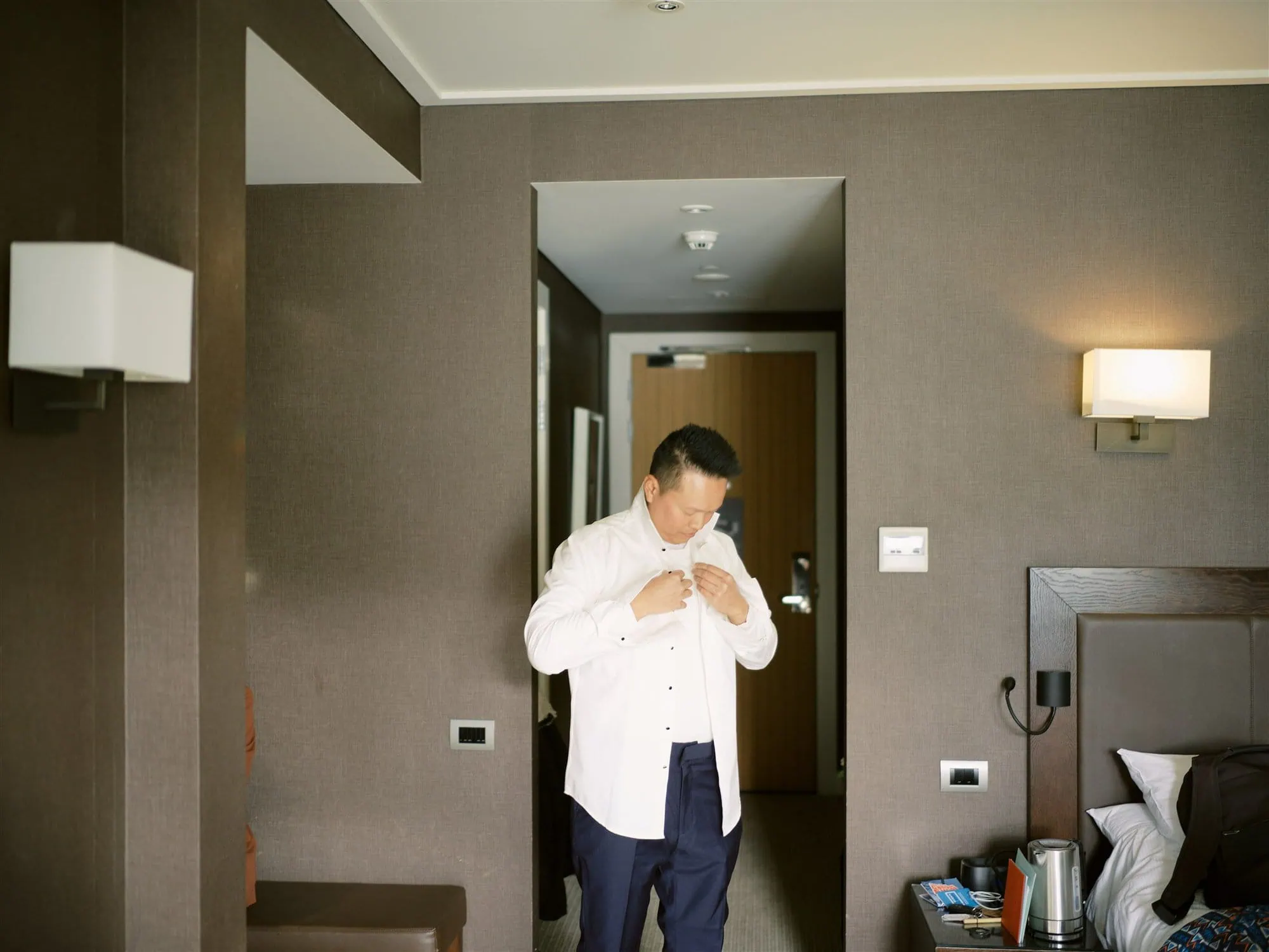 queenstown new zealand heli wedding elopement photographer videographer | In a hotel room, a man stands meticulously fastening his white shirt. Behind him, the calming ambiance is set by a neatly arranged bed, softly glowing wall lamps, and a table with a kettle and cups—a scene so perfect it feels like an auto draft of tranquility.