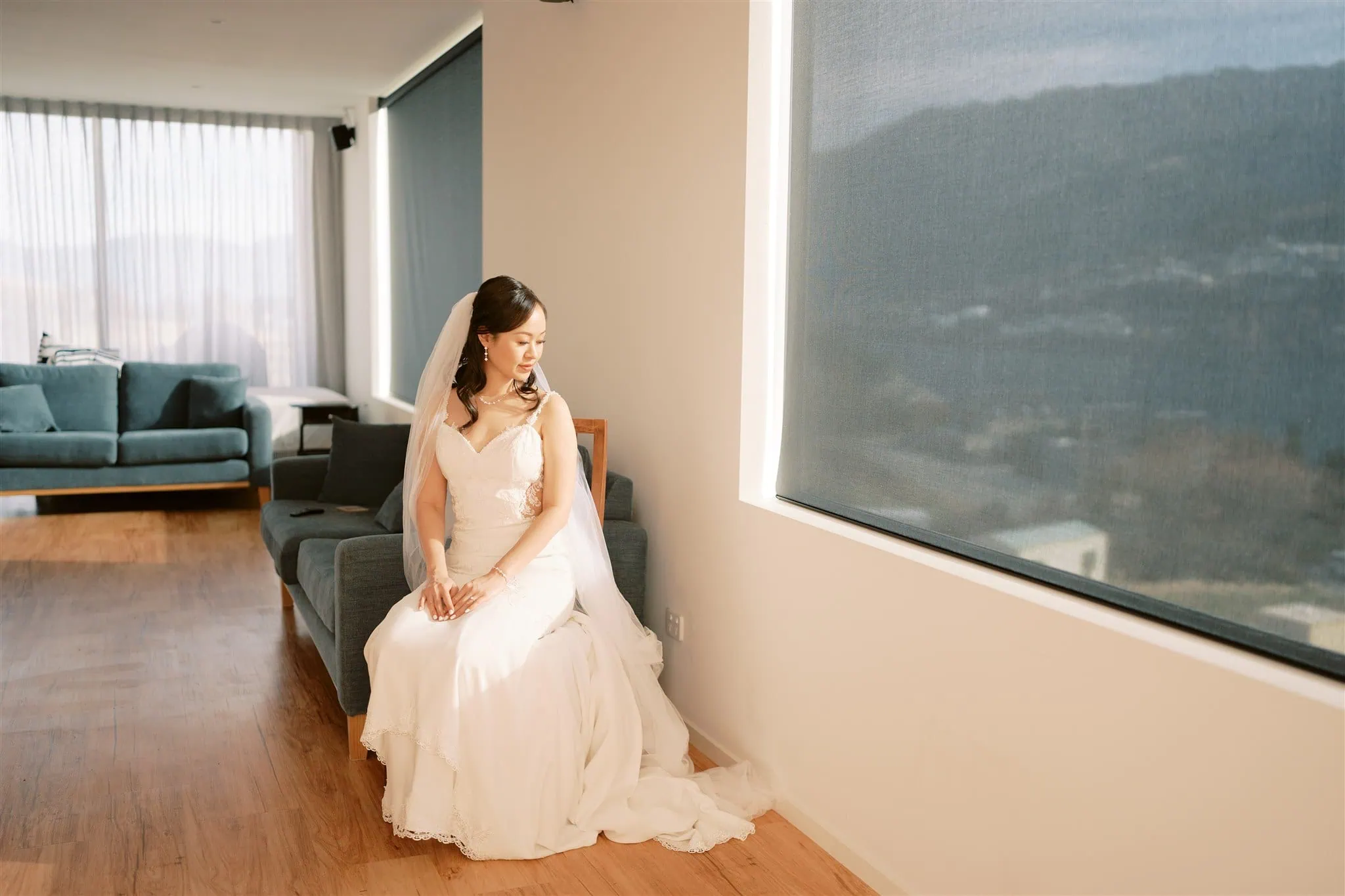 queenstown new zealand heli wedding elopement photographer videographer | A bride in a white dress sits on a sofa in a bright room, large windows with blinds casting soft light—a perfect moment of Queenstown wedding photography.