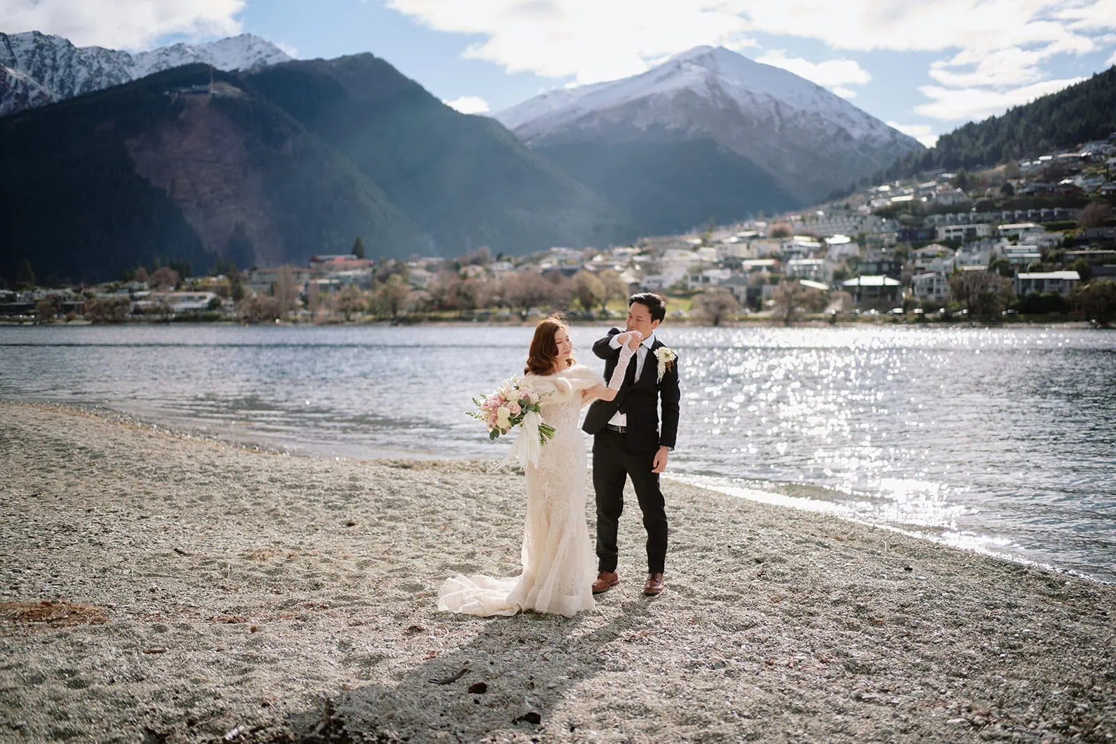 queenstown new zealand heli wedding elopement photographer videographer | In this stunning Queenstown pre-wedding photography shot, Ryan and Crystal stand elegantly on a lakeside beach, framed by snow-capped mountains and the quaint town in the background.